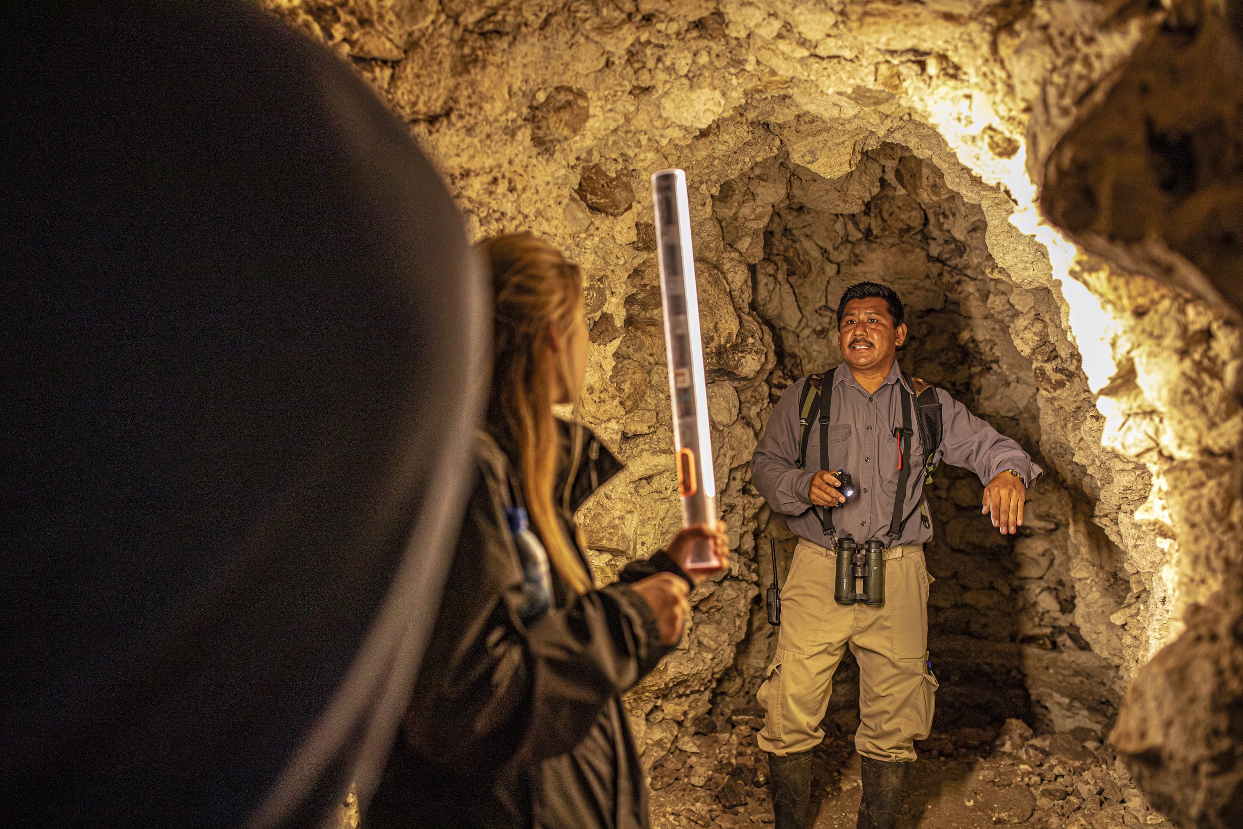 Guide speaking to tourists in a cave with rocky walls, holding a flashlight, with binoculars hanging around his neck.