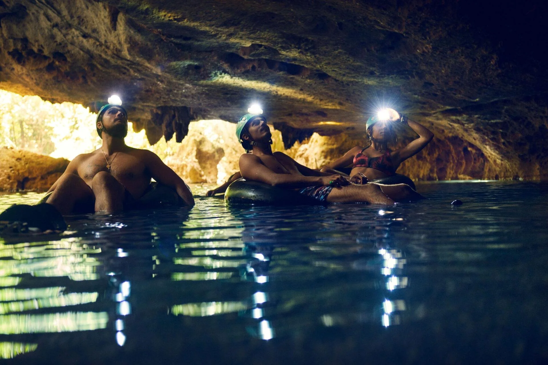 Three people with headlamps exploring inside a cave, sitting on floating tubes in a water-filled cavern.