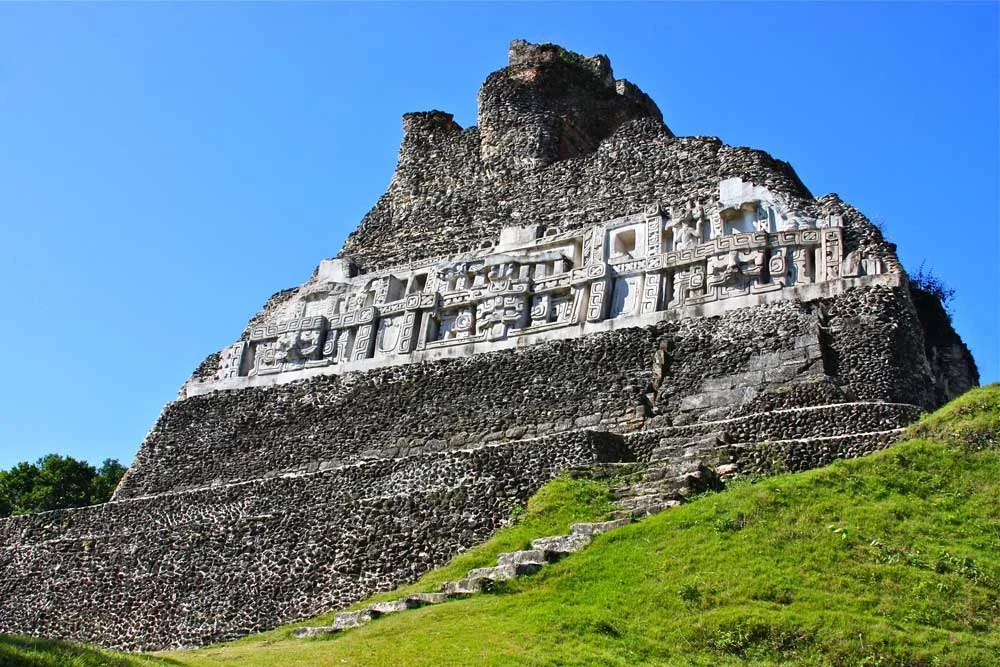 Ancient stone pyramid with intricate stone carvings near the top, set against a clear blue sky and green grass at the base.