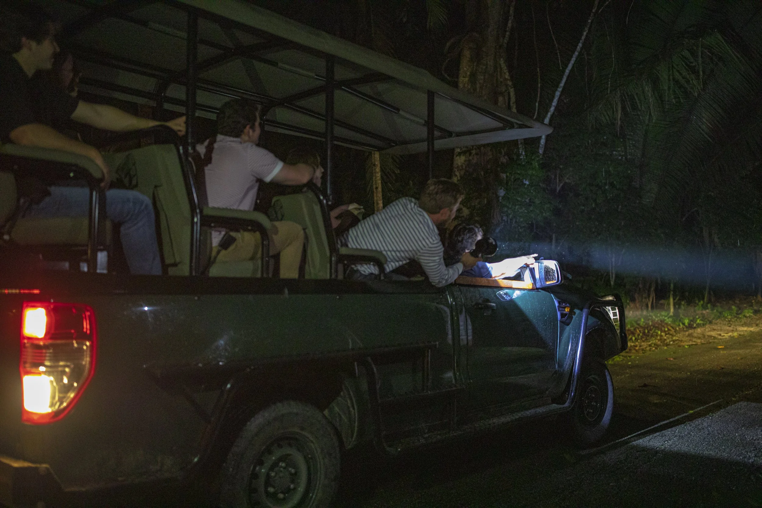 People on a night drive in the roofed back of a green pickup truck, one holding a flashlight