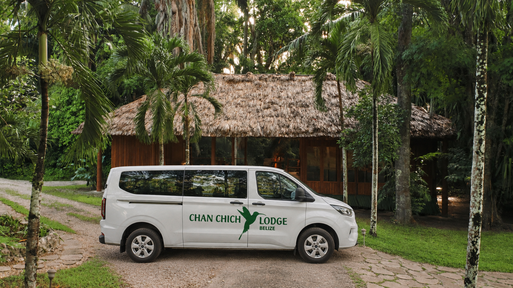 White van with 'Chan Chich Lodge Belize' written on the side parked on a gravel driveway in front of a thatched roof lodge surrounded by lush tropical trees.