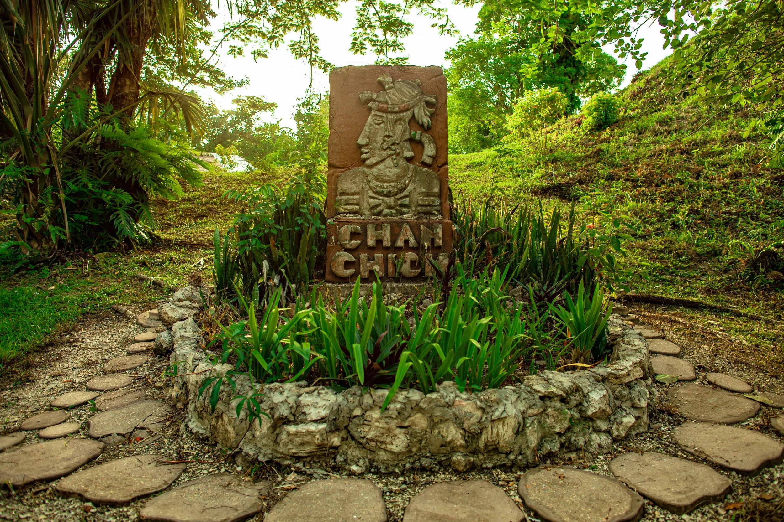Carved stone sign reading Chan Chich, with a carved figure in a headdress, set in a ring of stones in the garden