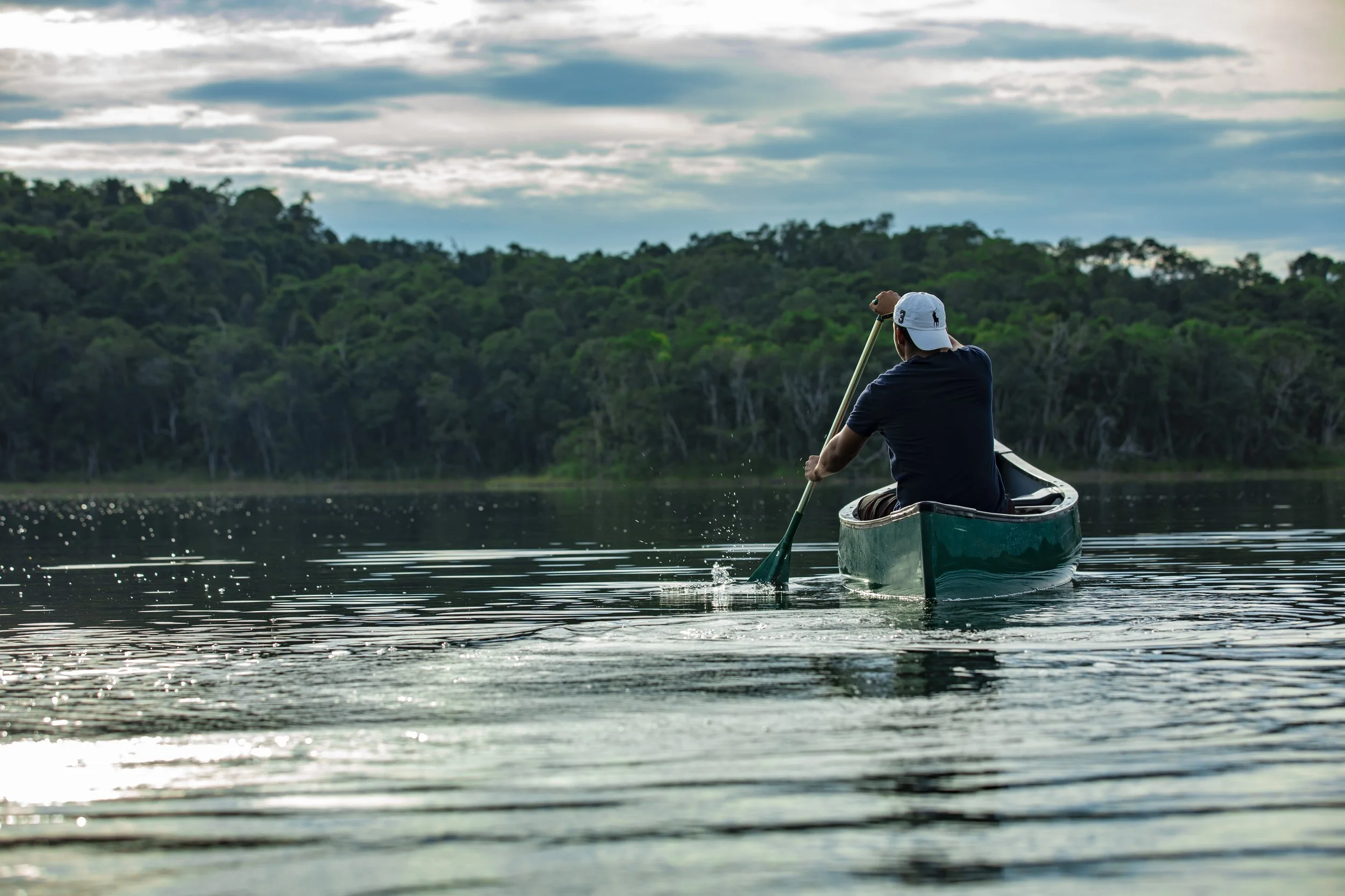 Person paddling a canoe on a calm lagoon, with dense green rainforest and a cloudy sky behind