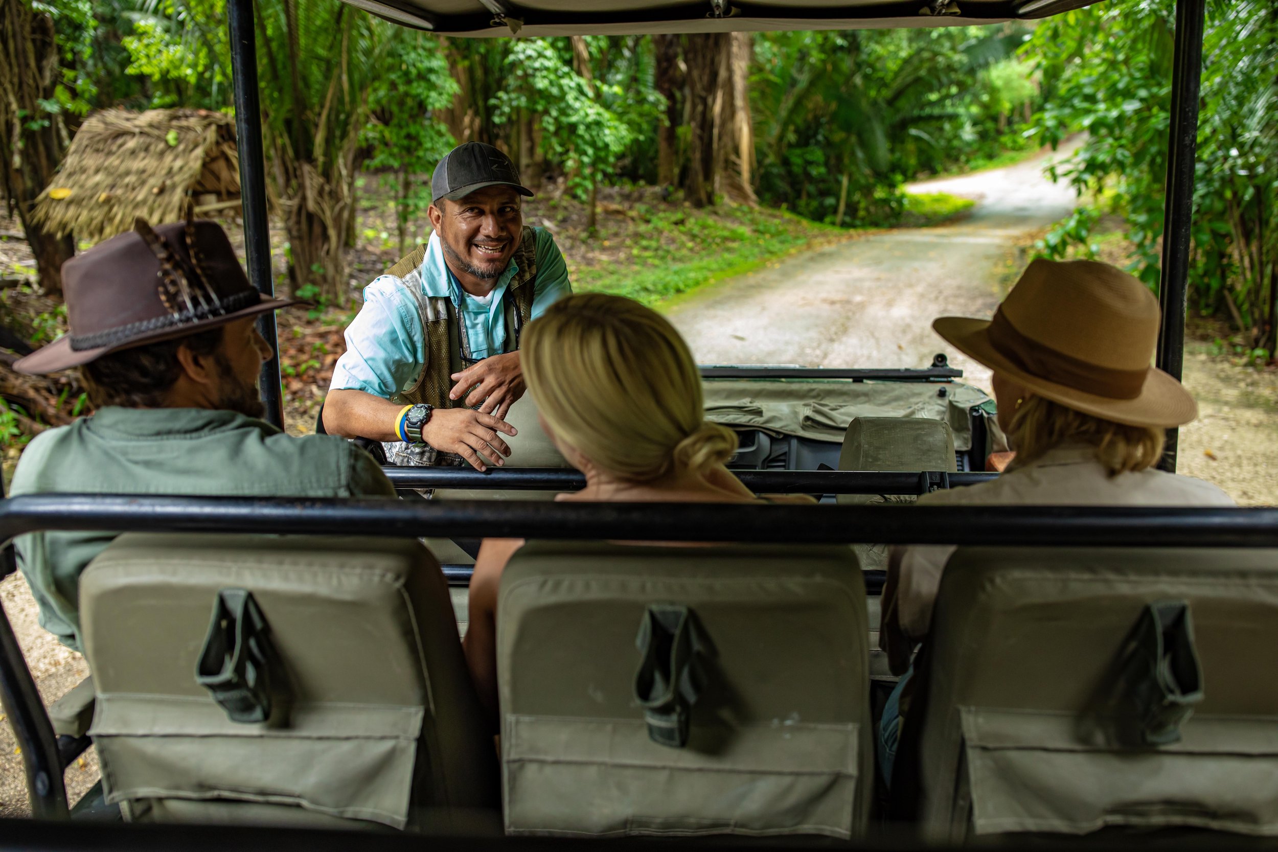 A guide turns to talk with guests seated in an open safari vehicle on a dirt road