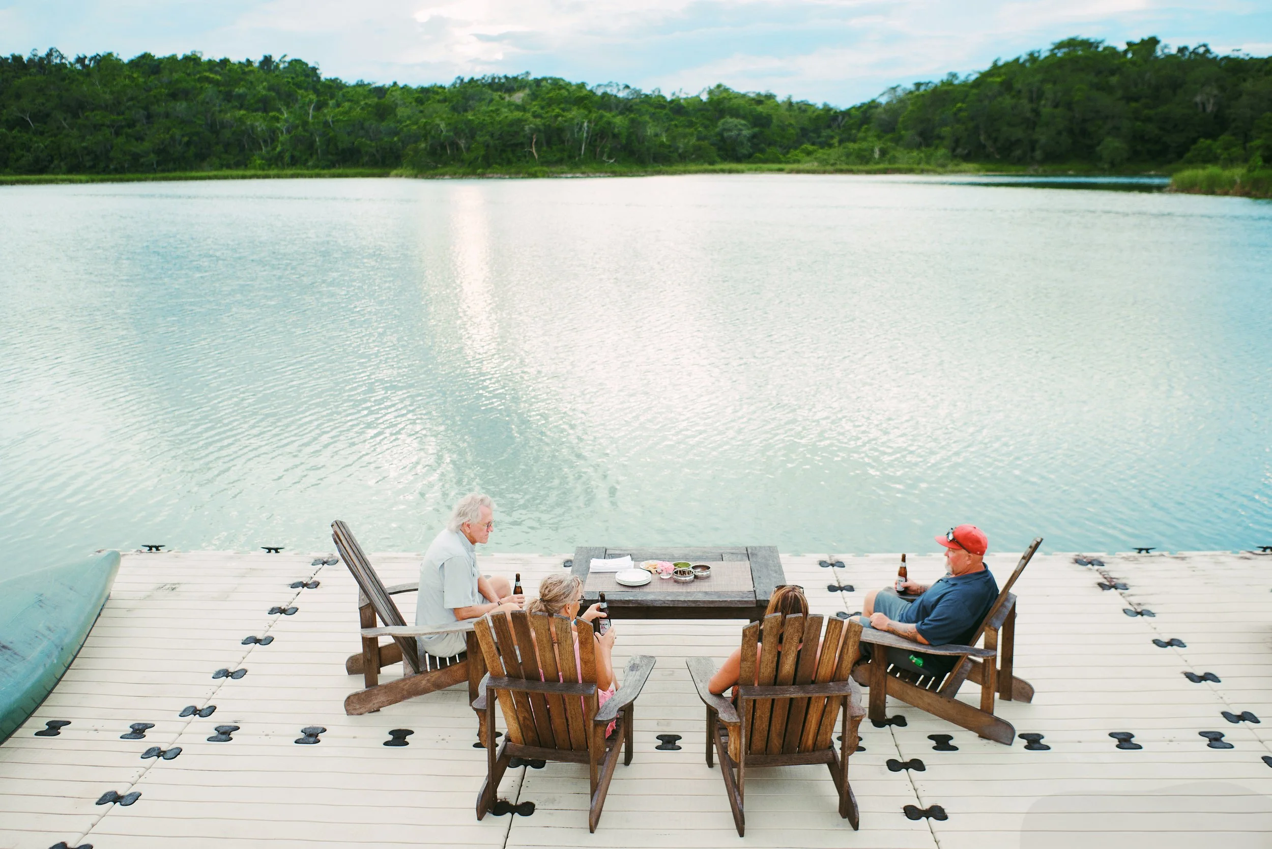 People sitting at a table on a dock by a large body of water with trees in the background.