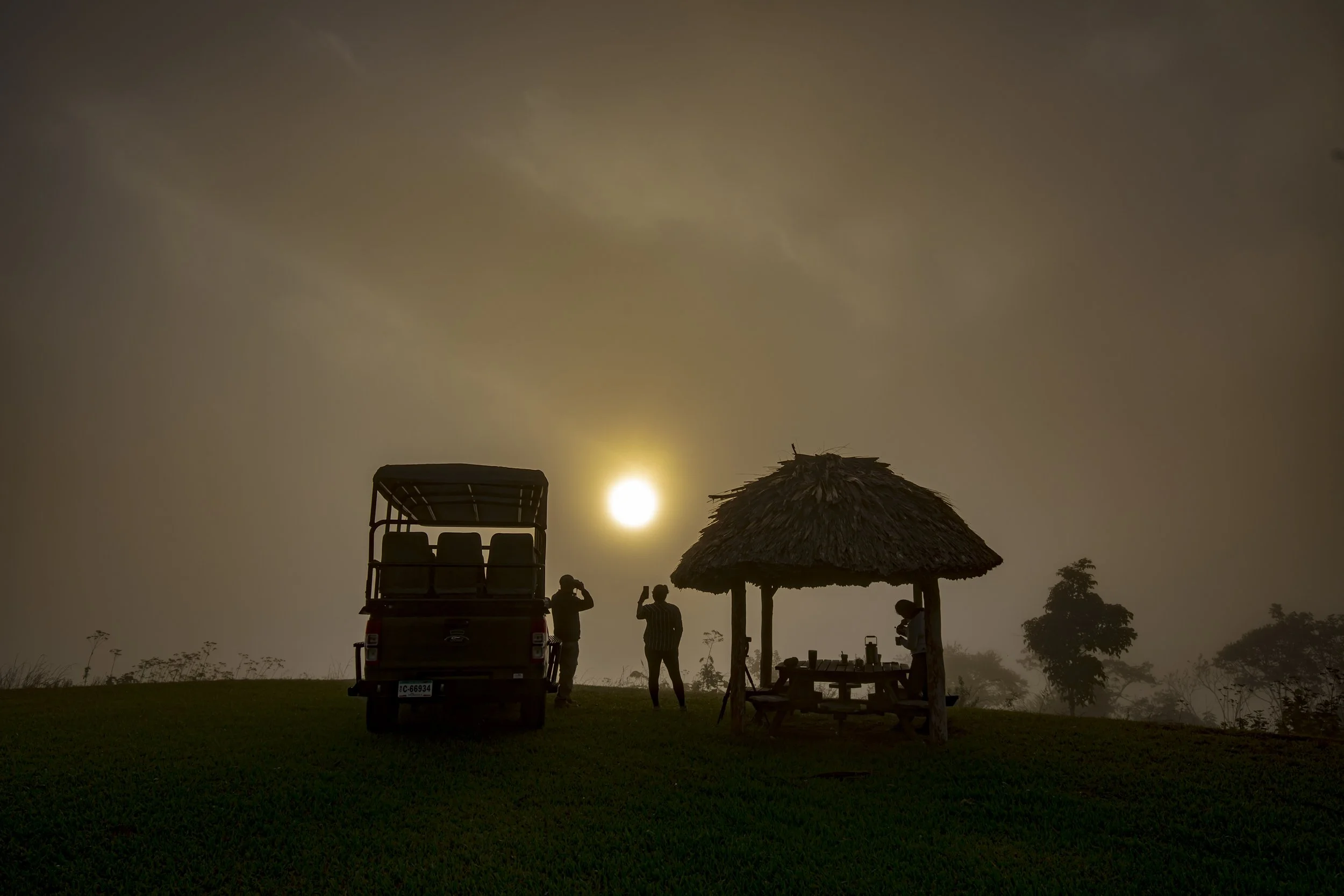 People in silhouette under a thatched shelter at sunset, with a vehicle parked nearby