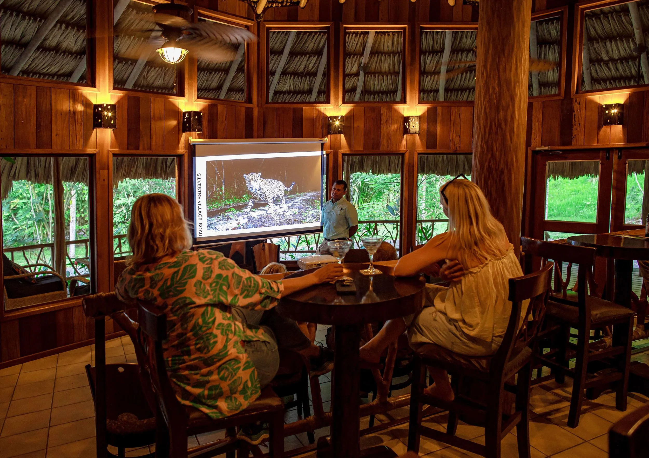 Guests at tables in a wood-and-thatch room watching footage of a spotted cat on a large screen