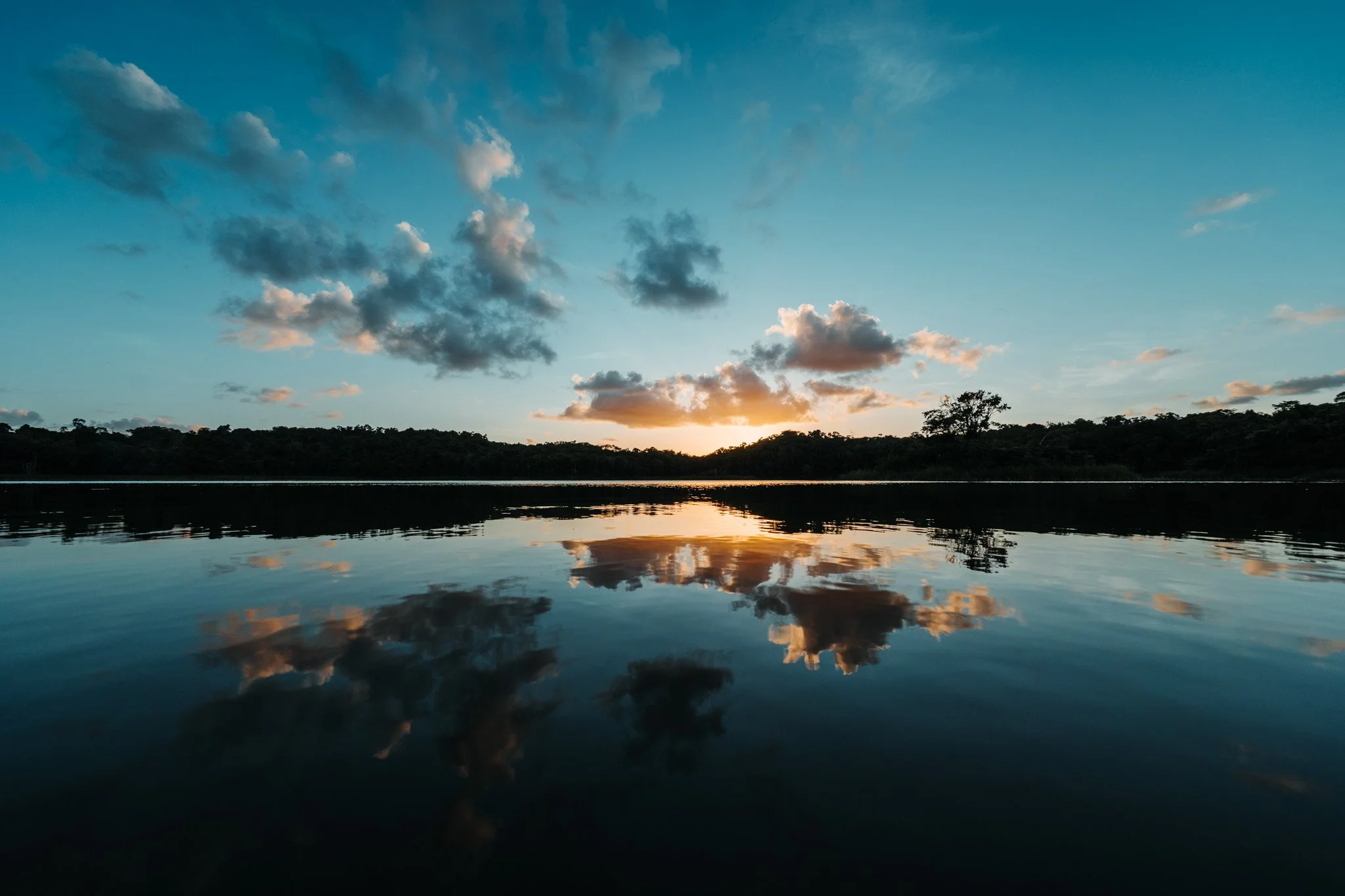 Sunset over a calm lake, the tree line dark on the horizon and the sky reflected in the water