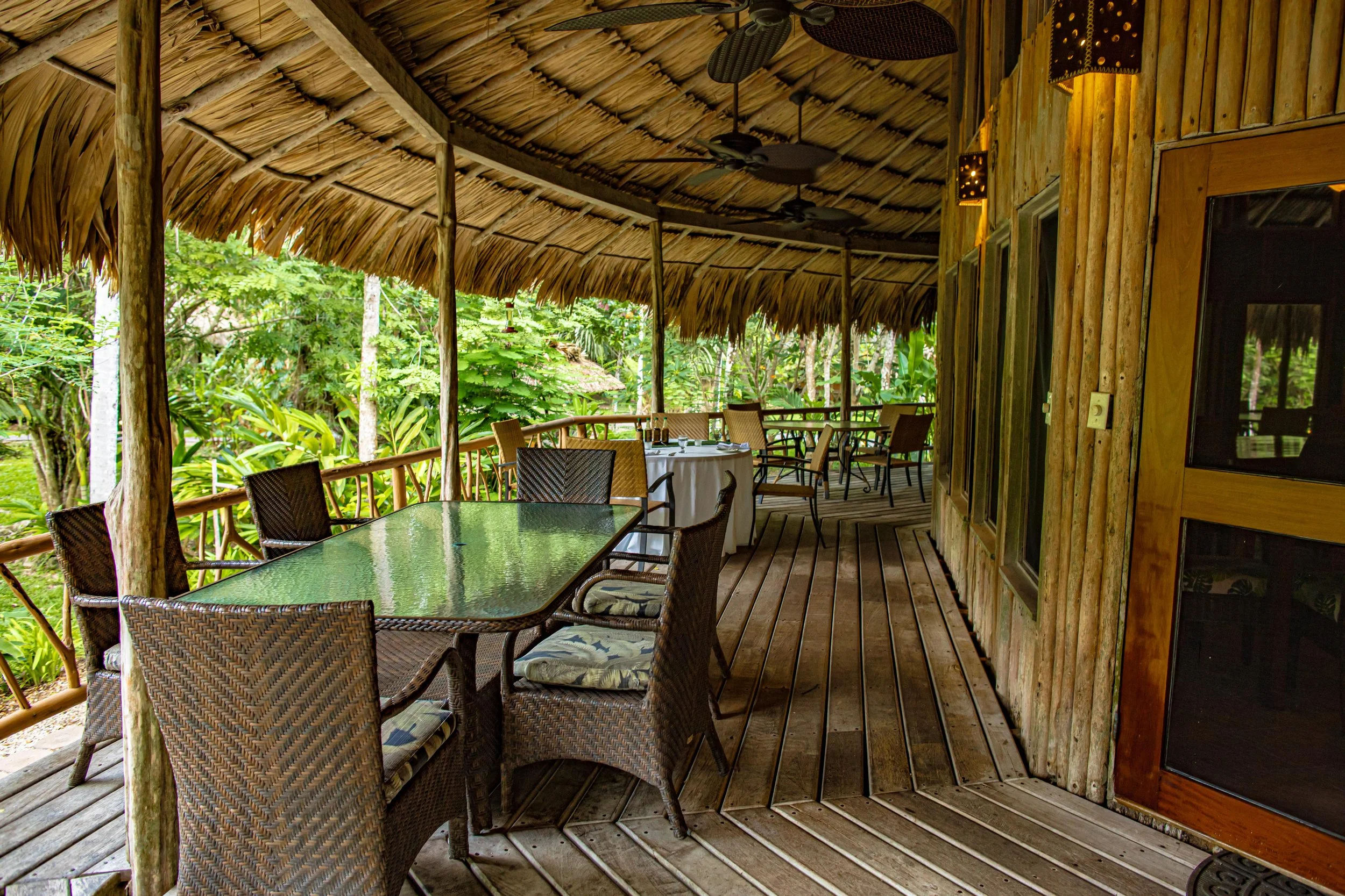 Open-air patio under a thatched roof with bamboo walls, wicker chairs and a glass-topped table among tropical plants