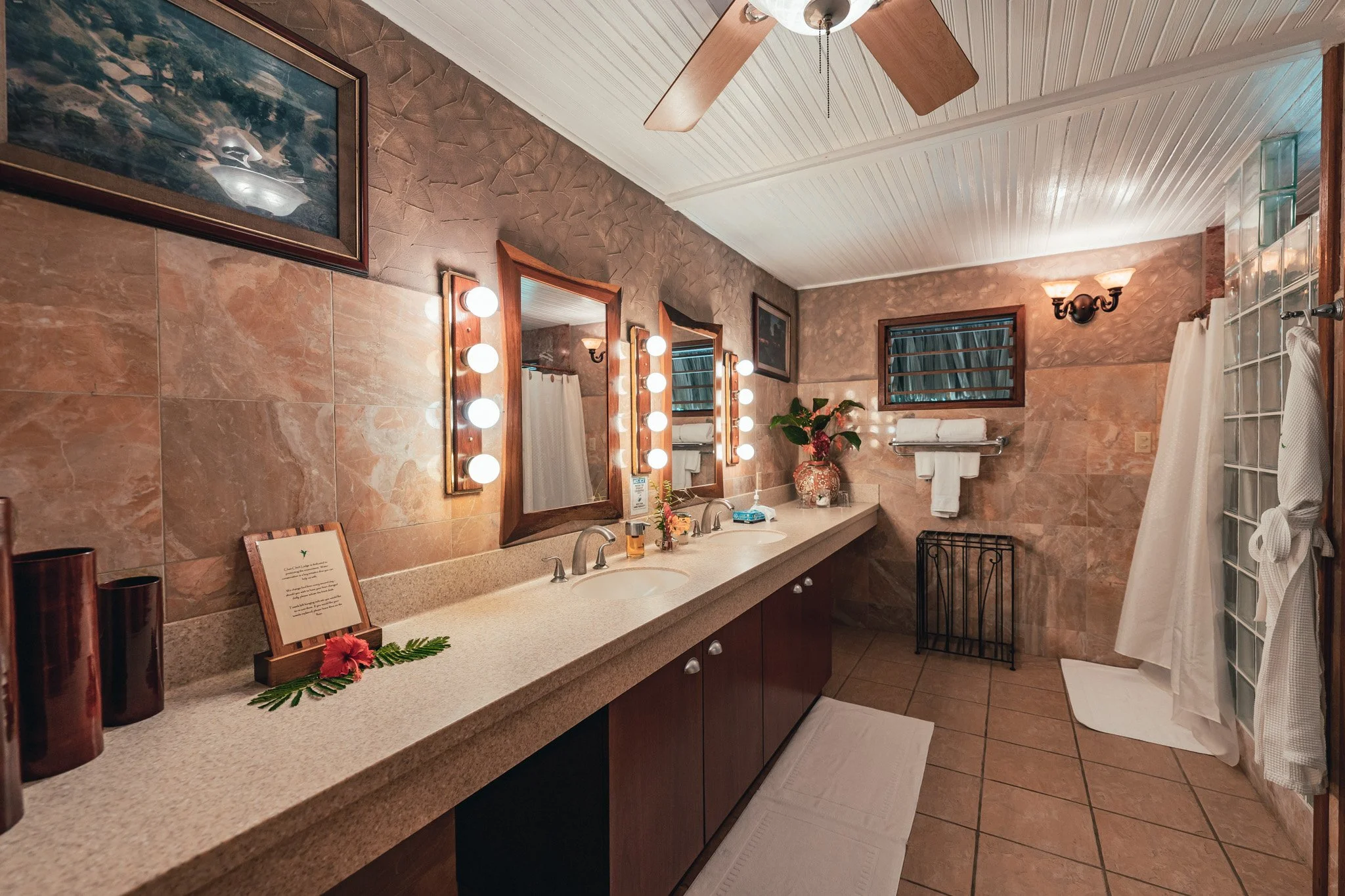 Bathroom with a double-sink vanity, lighted mirrors, framed art, flowers on the counter and towels folded on a rack
