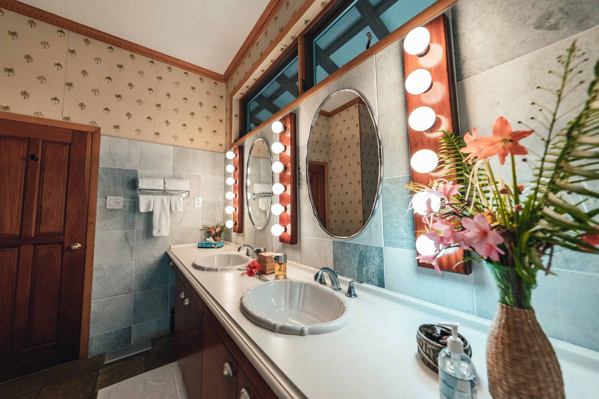 Bathroom with a double-sink vanity, wood-framed mirrors with white globe lights and palm-print wallpaper