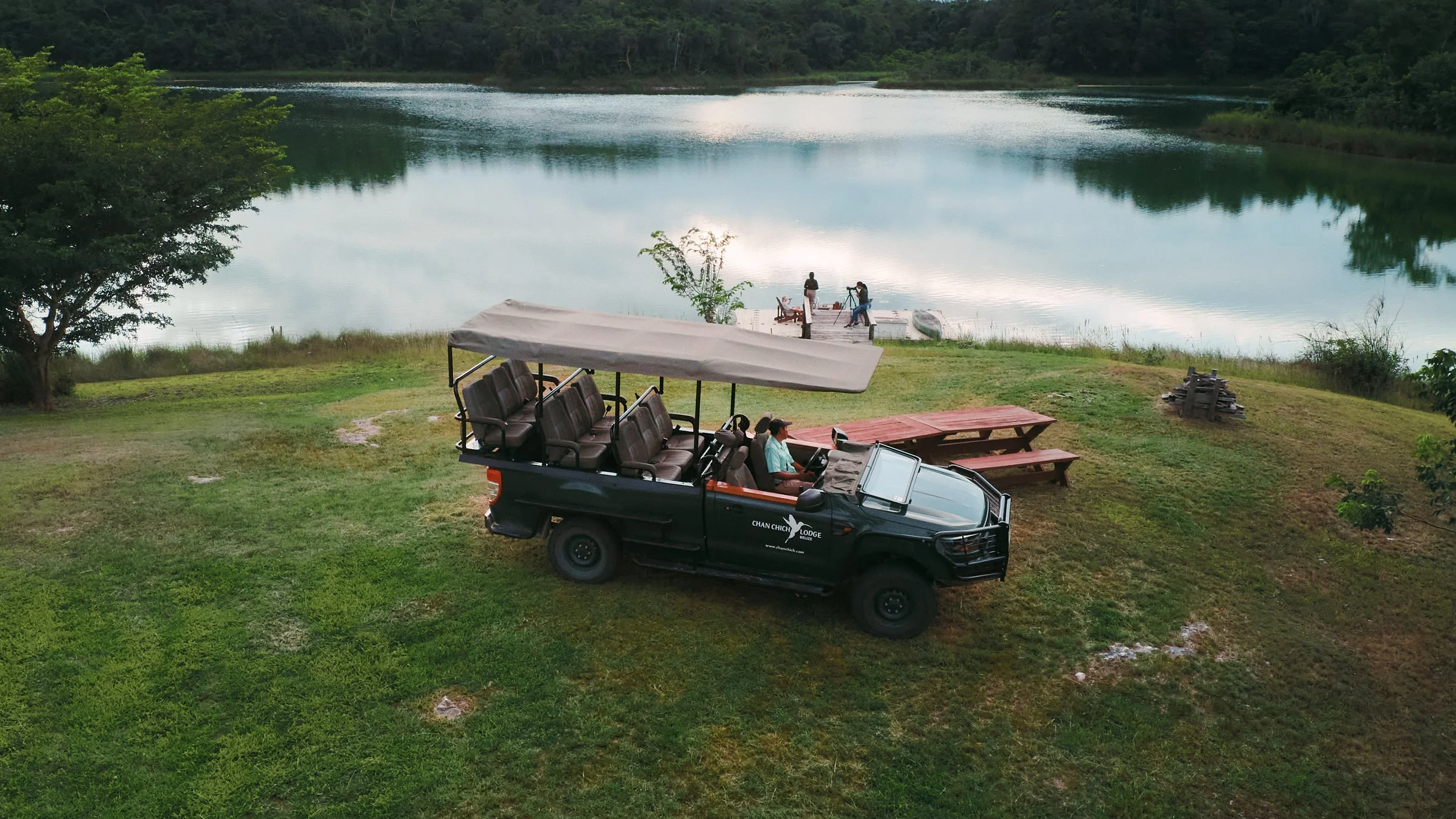 Open safari vehicle parked on the grass above a lagoon, with people on the dock below