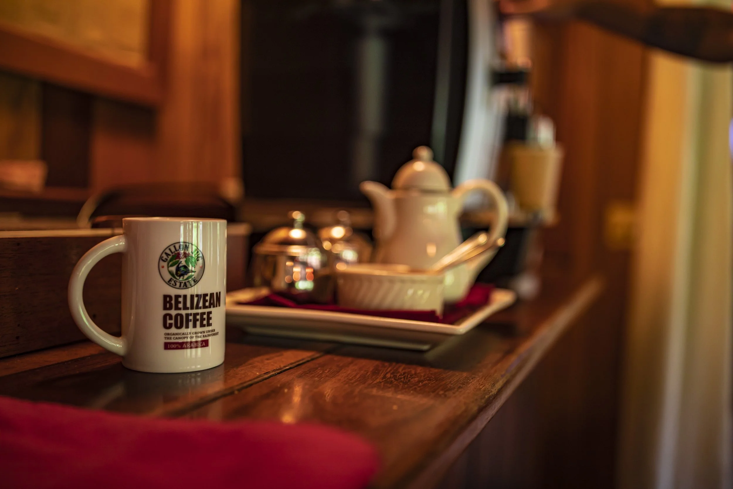 White mug printed with the words Belizean Coffee on a wooden counter, with teapots and a tray behind it