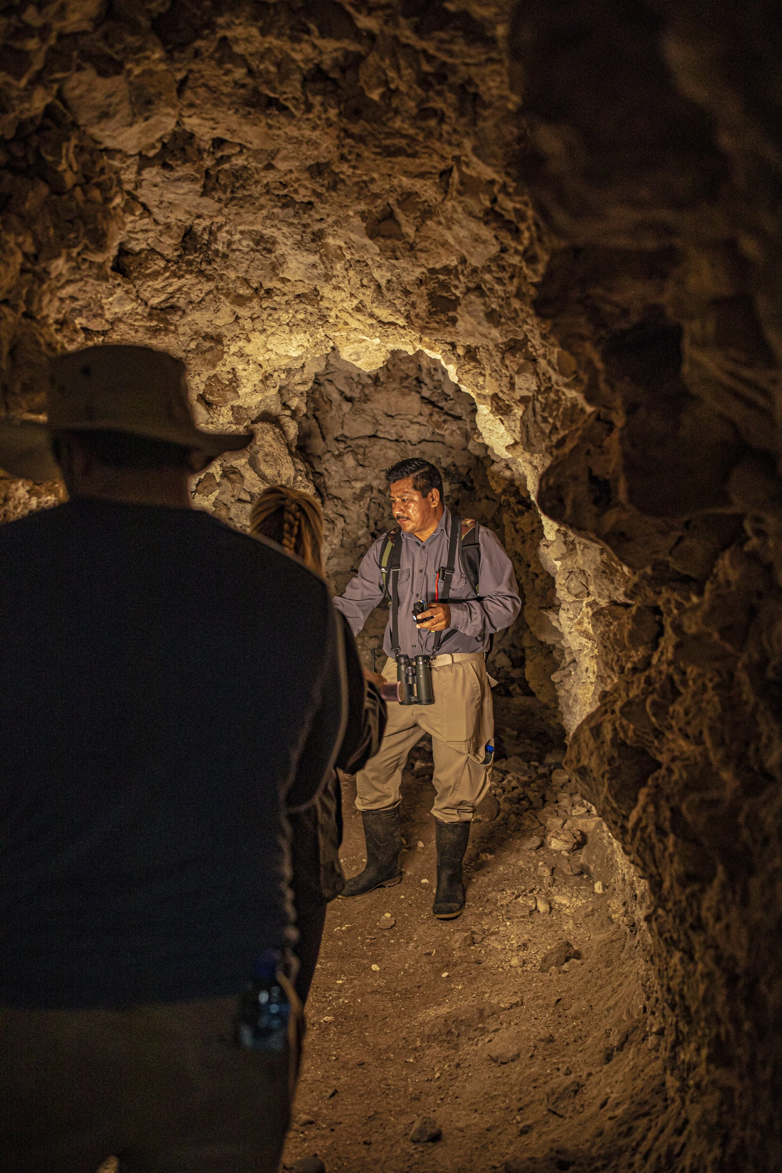 A man wearing tan pants, black boots, and a backpack, holding a camera, standing inside a rocky cave while talking to a group of people.