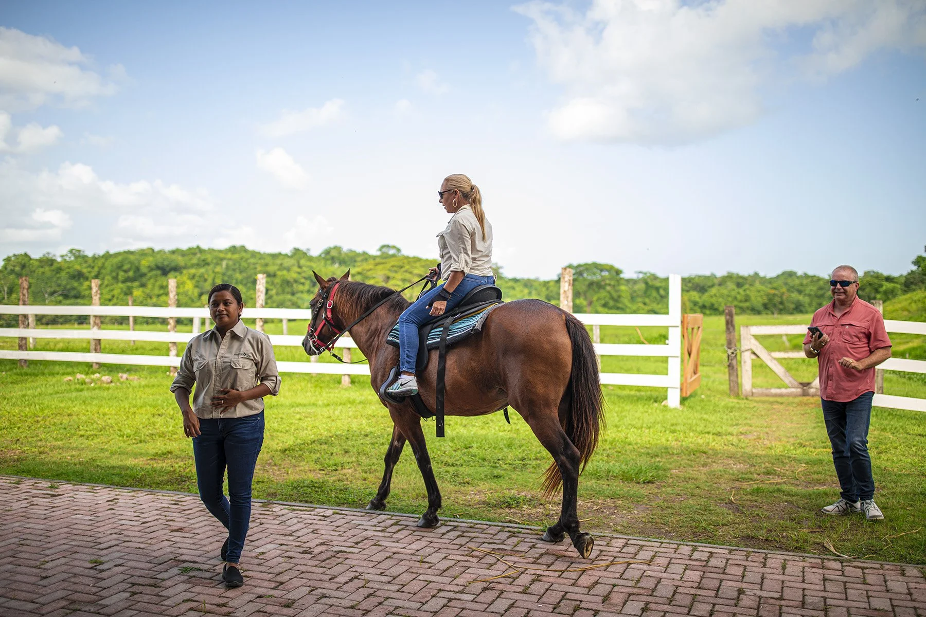 A woman is riding a brown horse in an outdoor area surrounded by a white fence, with two assistants nearby, one on each side of the horse, and a man standing with a phone in hand. The background features green trees and a partly cloudy sky.