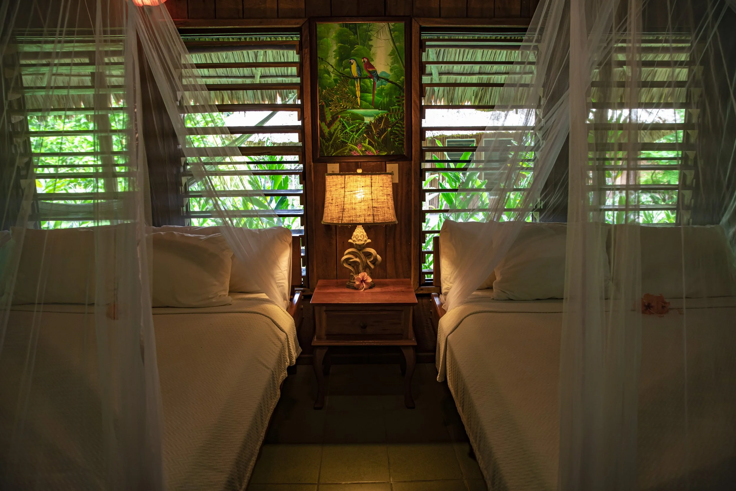 View of a bedroom with two twin beds, a wooden nightstand, and a table lamp in the center. Behind the beds are wooden slat windows with sheer curtains, revealing lush greenery and a painting of colorful parrots in a jungle scene on the wall.