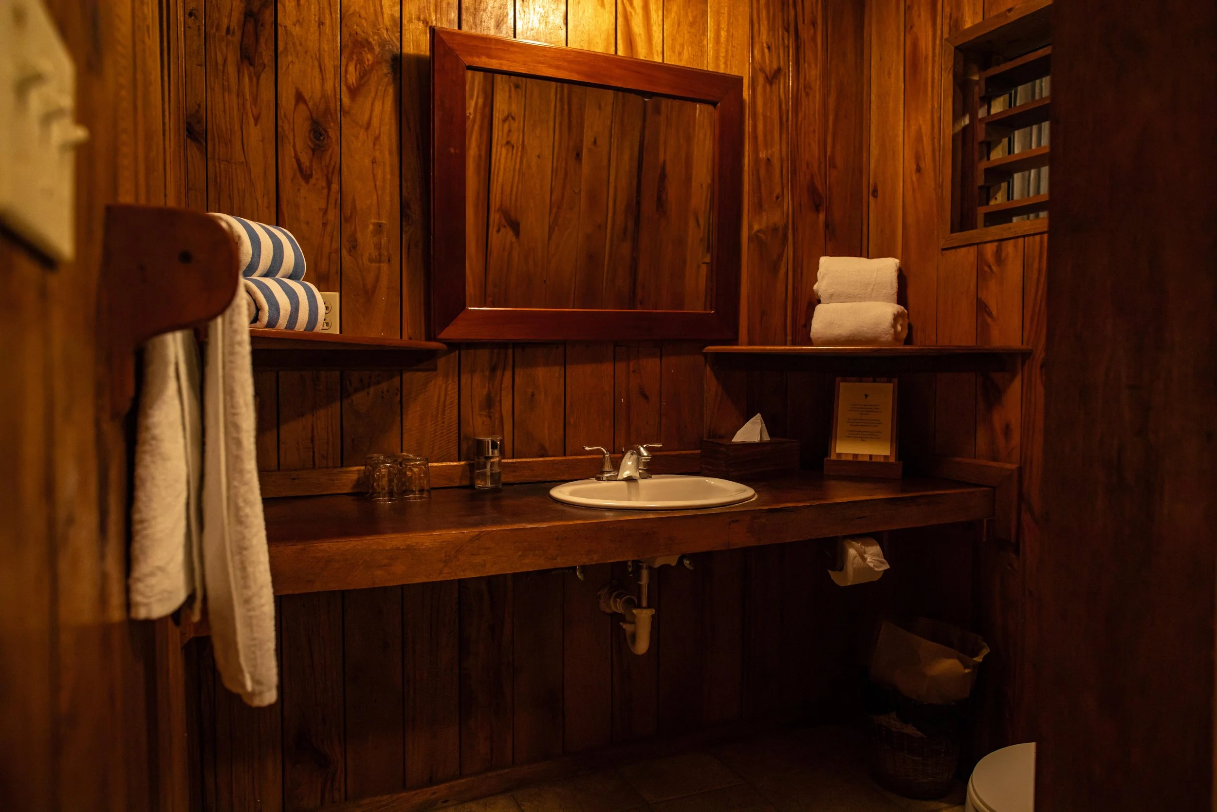 Wood-paneled bathroom with a small sink, countertop, mirror, shelves with towels, and a window.