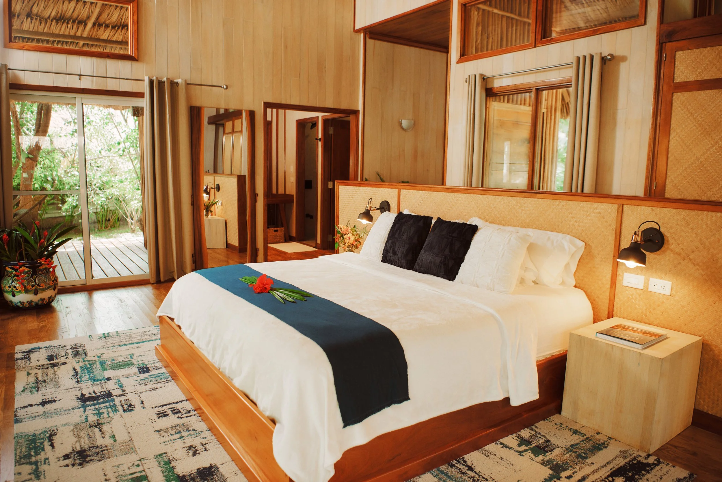 Bedroom with wooden walls, a large bed in white linens, a bedside table with a book, and a glass door onto a deck