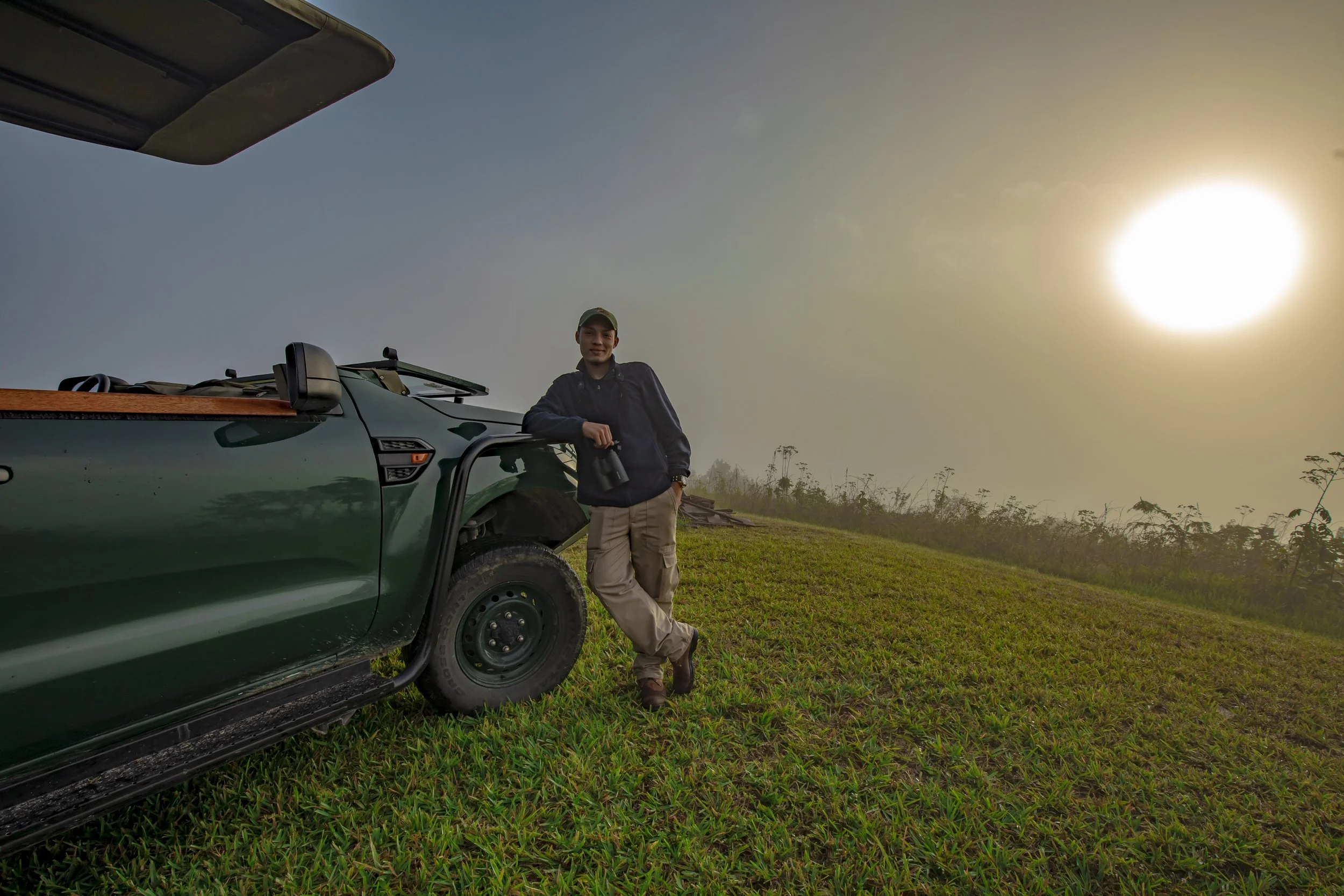 Man standing beside a green pickup truck on the grassy top of the Escarpment as the sun sits low