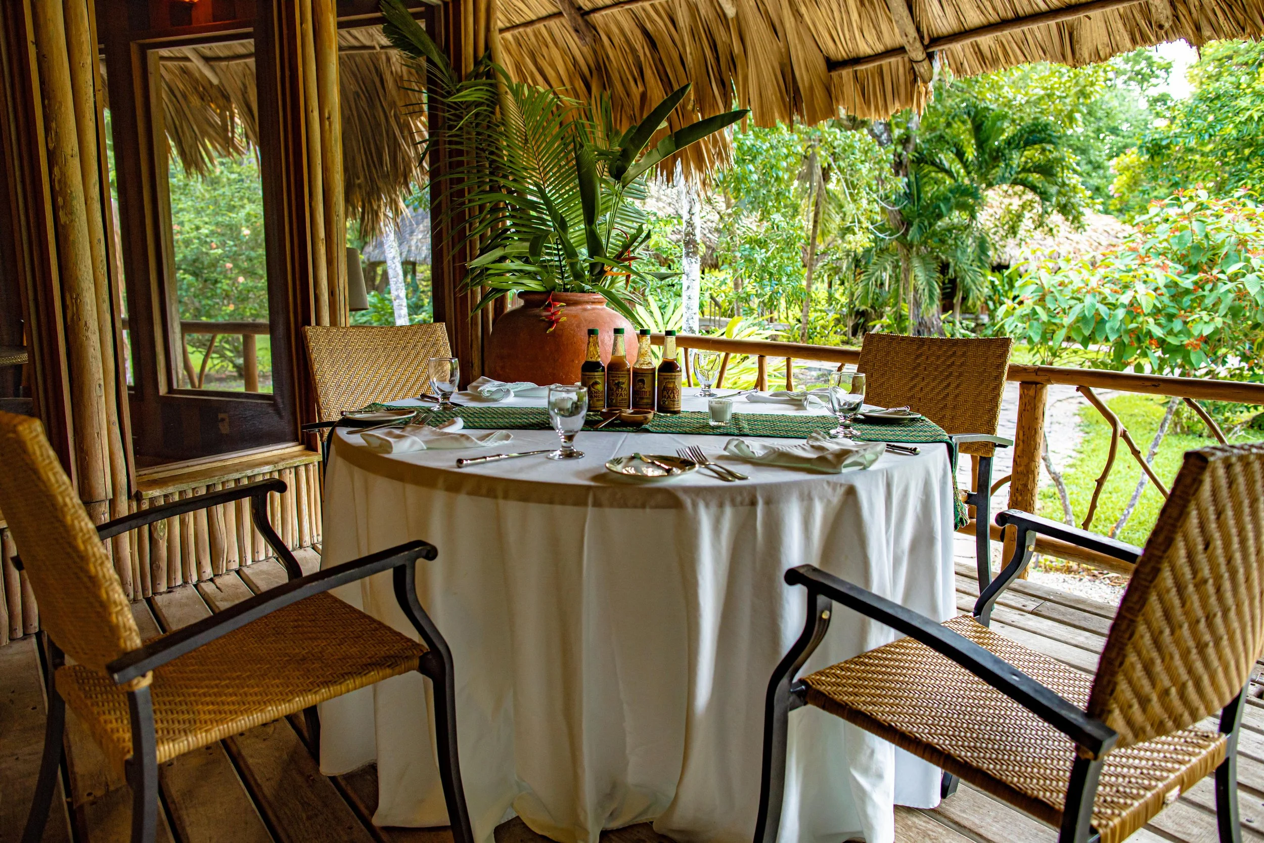 Round table set with a white cloth, wine glasses and cutlery, with wicker chairs, outdoors among green plants