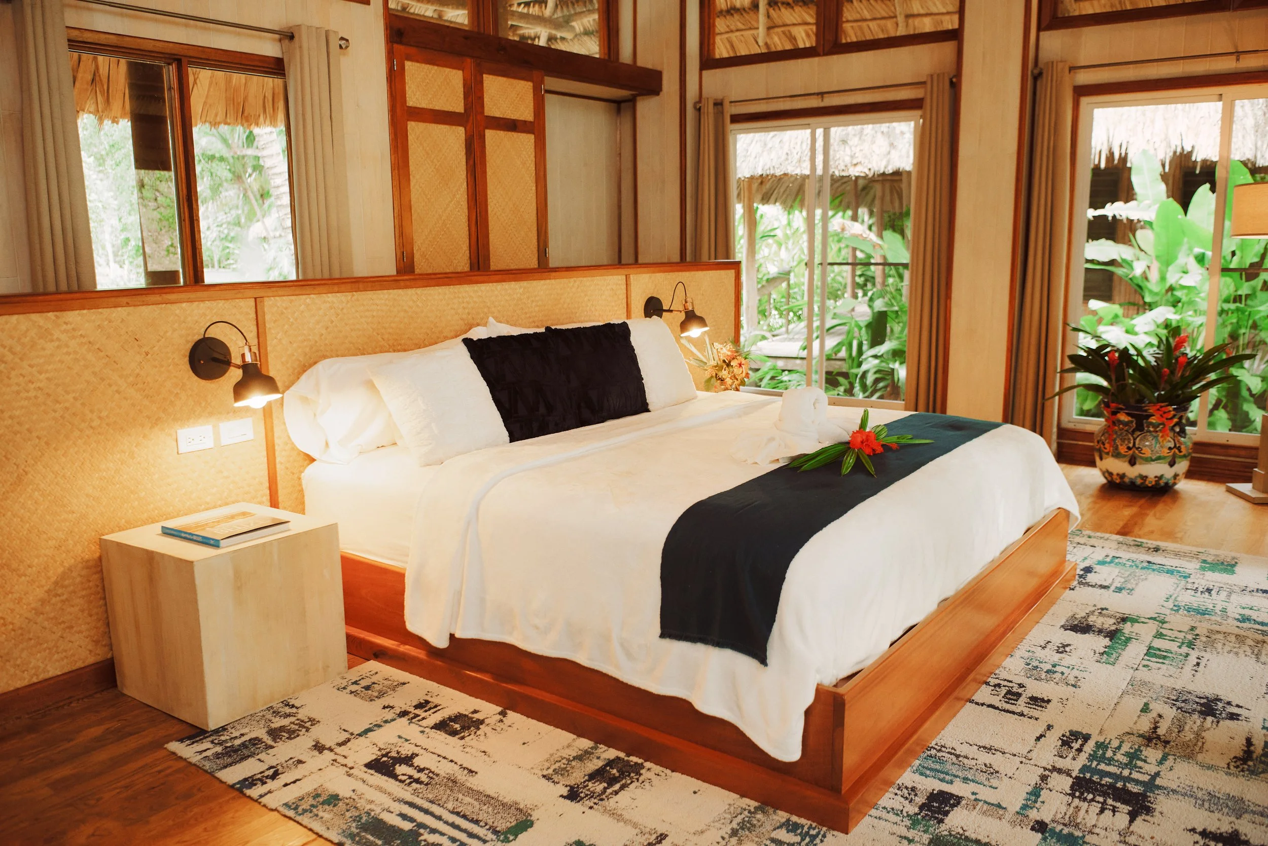 Bedroom with a large bed in white linens with black pillows and throw, a nightstand with a book and lamp, and a potted plant