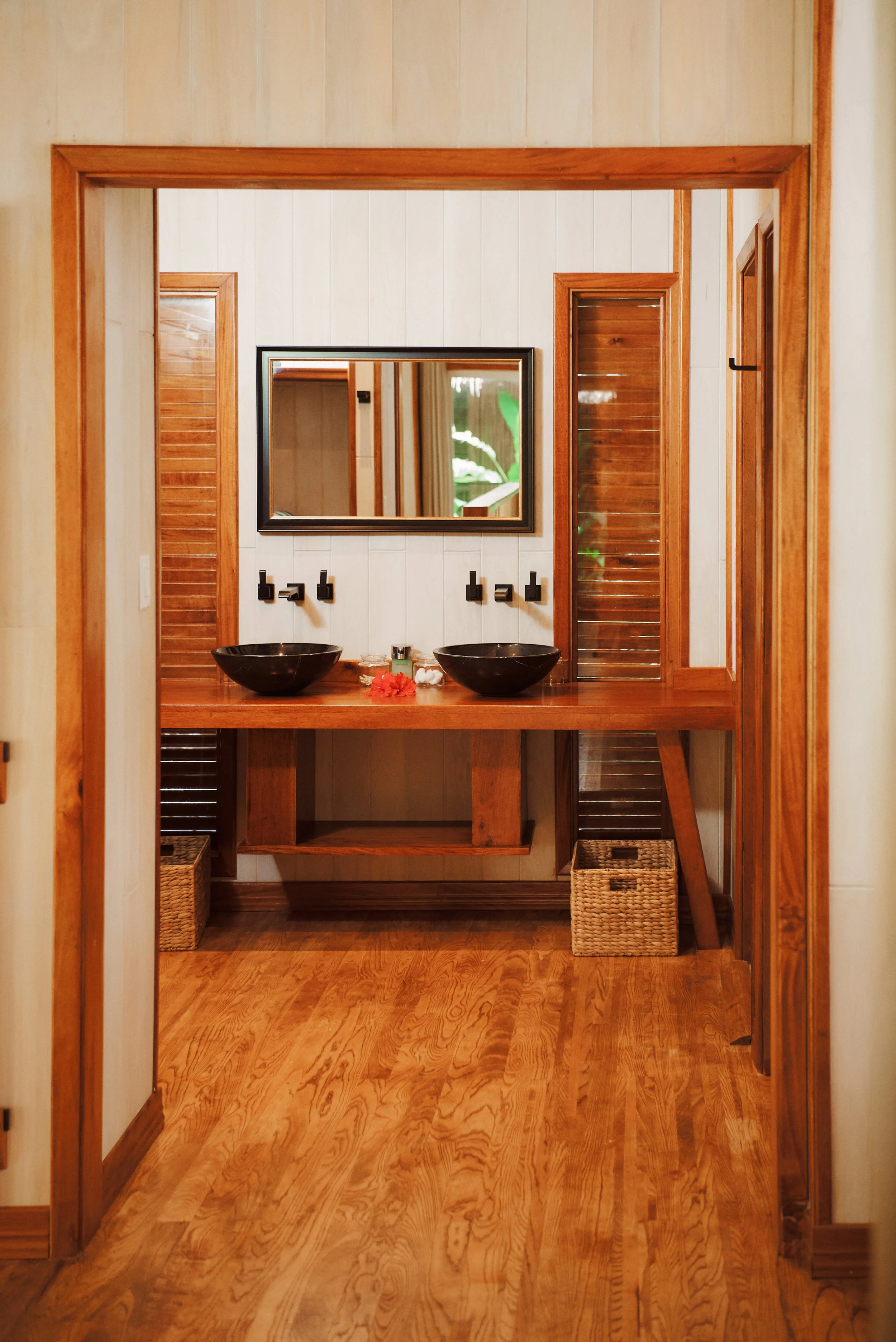 A bathroom vanity with two black vessel sinks, a mirrored wall, wooden cabinets, and woven baskets on the floor.