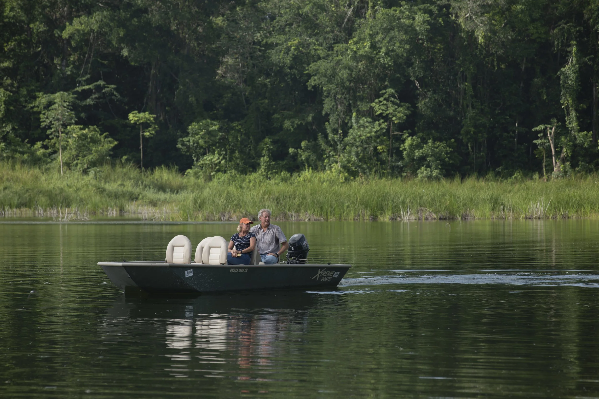 A small motorboat with two people, a man and a woman, floating on a calm lake surrounded by green trees and tall grass.