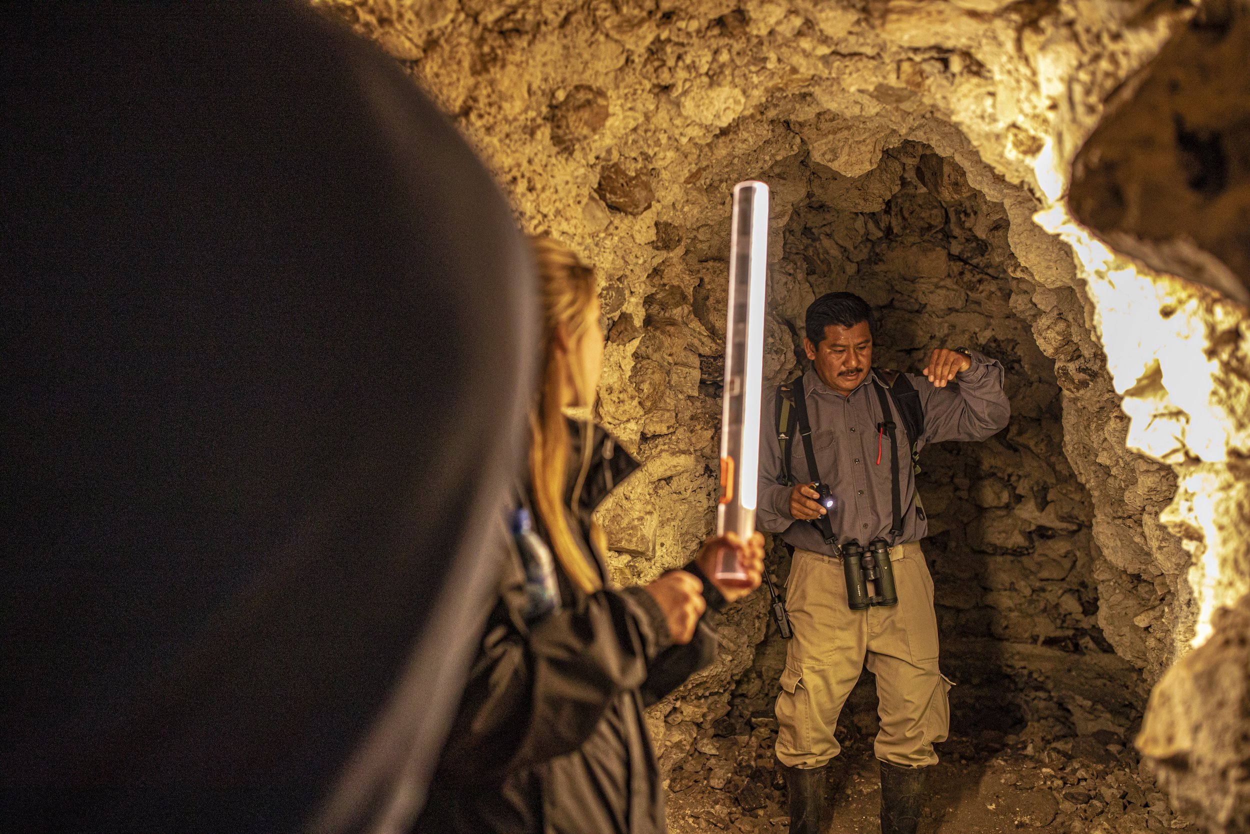 Two people with a flashlight and binoculars inside a rocky underground passage lit by warm light