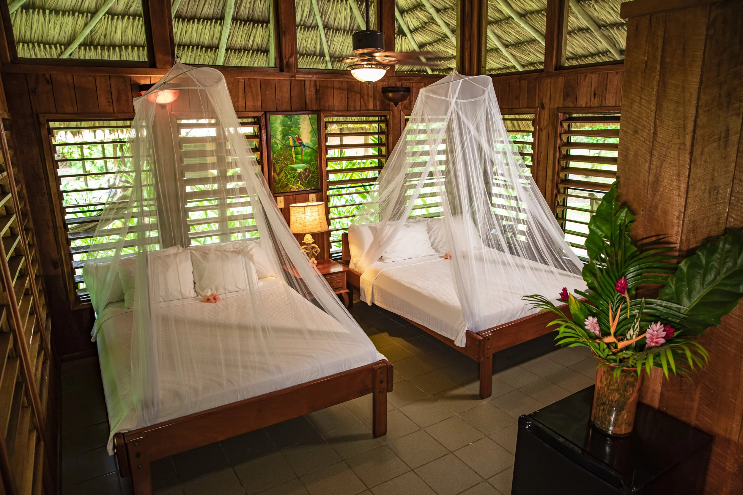Two beds under mosquito nets in a wood-walled room with louvered shutters and a vase of flowers