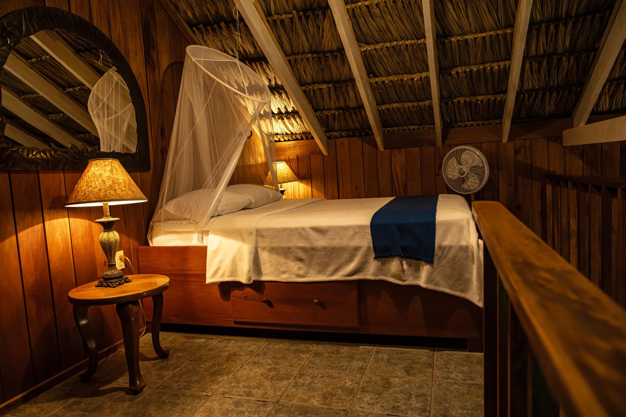 Wood-paneled bedroom with a bed under a mosquito net, bedside lamps, a mirror and a fan