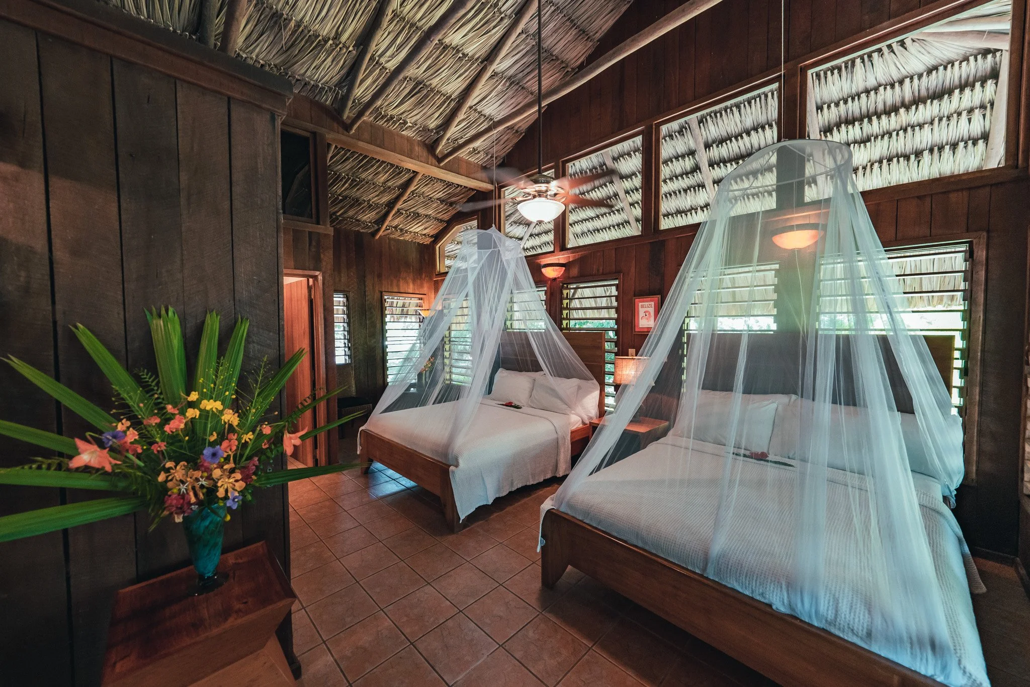 Two beds under mosquito nets in a room with wooden walls and ceiling, a ceiling fan and slatted wooden windows