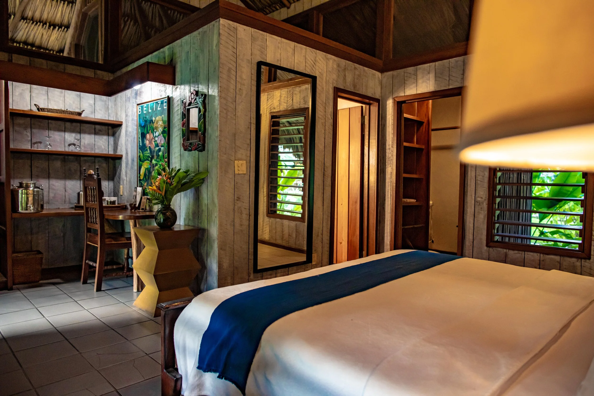 Wood-paneled bedroom with a large bed in white and blue, a tall mirror, a BELIZE sign, plants and shuttered windows
