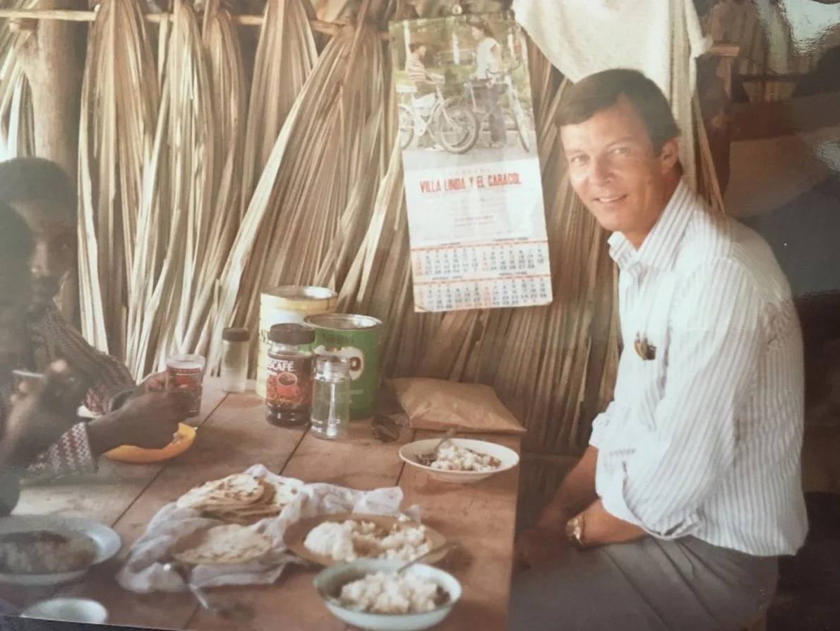 A man in a white shirt at a table set with plates of food, beneath a palm-thatch wall
