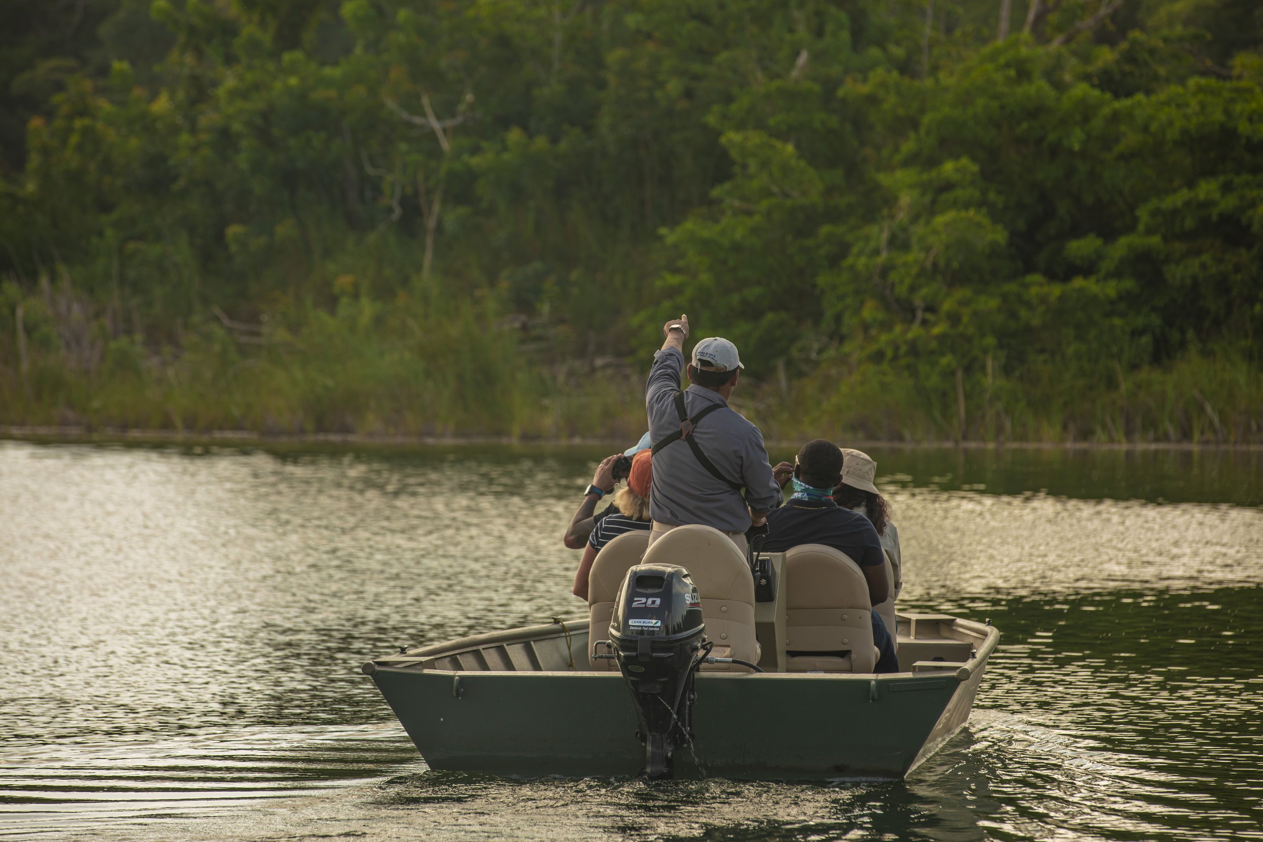 A group of people on a boat in a river, surrounded by green trees, with one person standing and pointing outward.