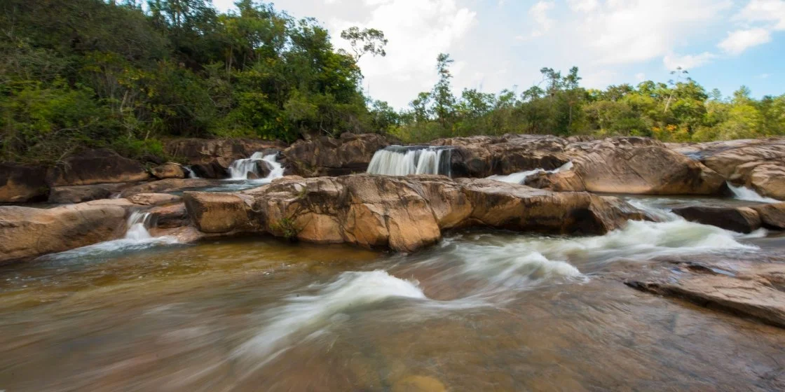 A flowing river with small waterfalls surrounded by rocks and lush green trees under a partly cloudy sky.