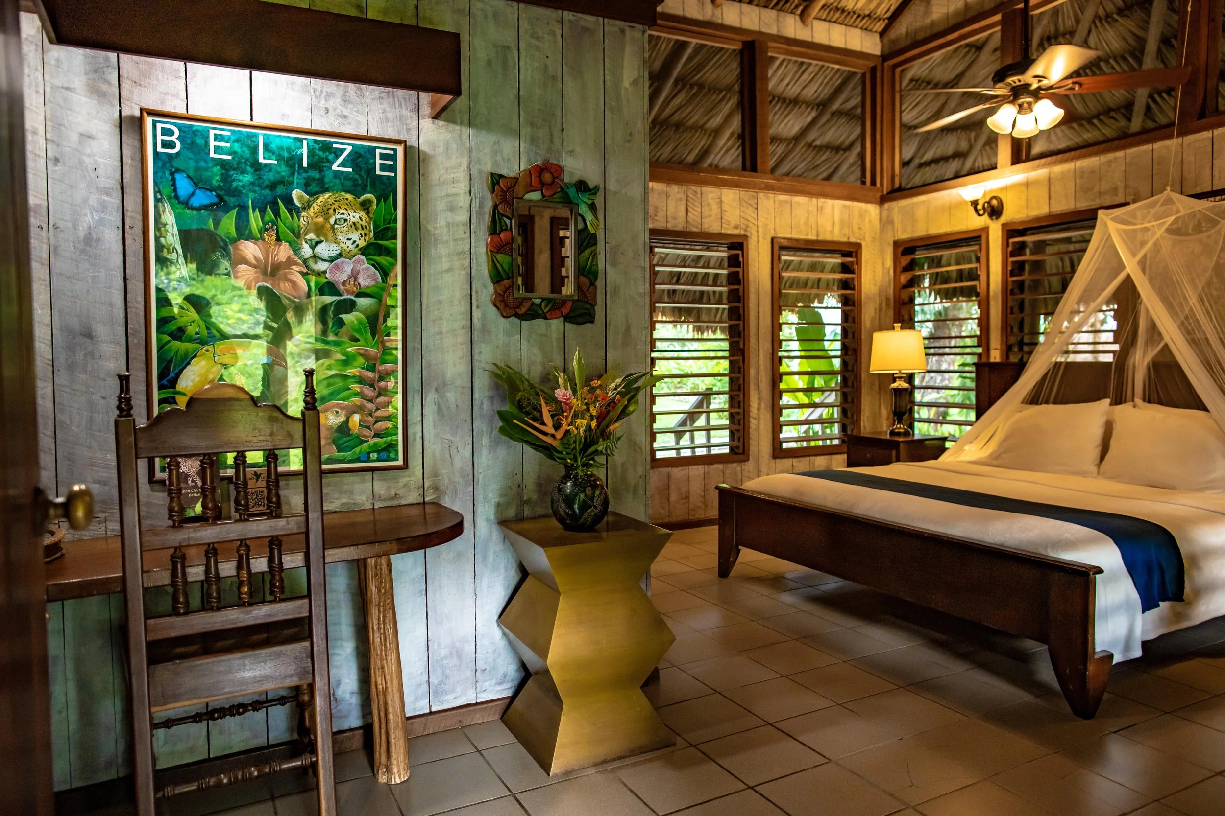 Bedroom under a thatched ceiling with a bed under a mosquito net, a bedside lamp, a mirror, a wildlife painting and flowers
