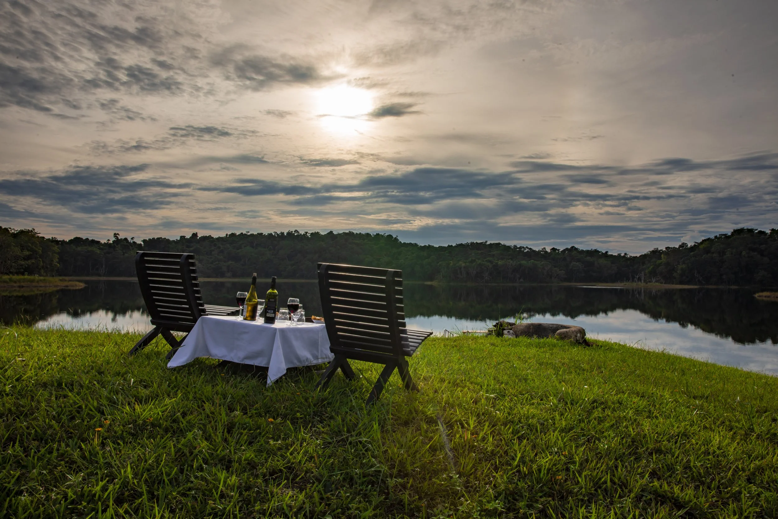 Two empty chairs facing a lake at sunset beside a table with wine and small plates