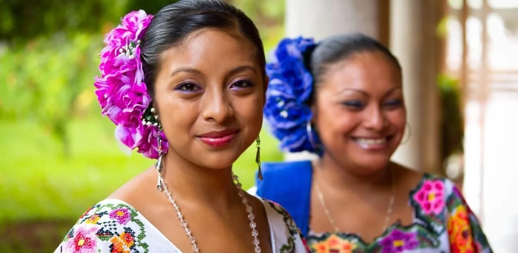 Two women dressed in traditional Mayan attire with colorful floral embroidery and large floral headpieces, smiling outdoors.