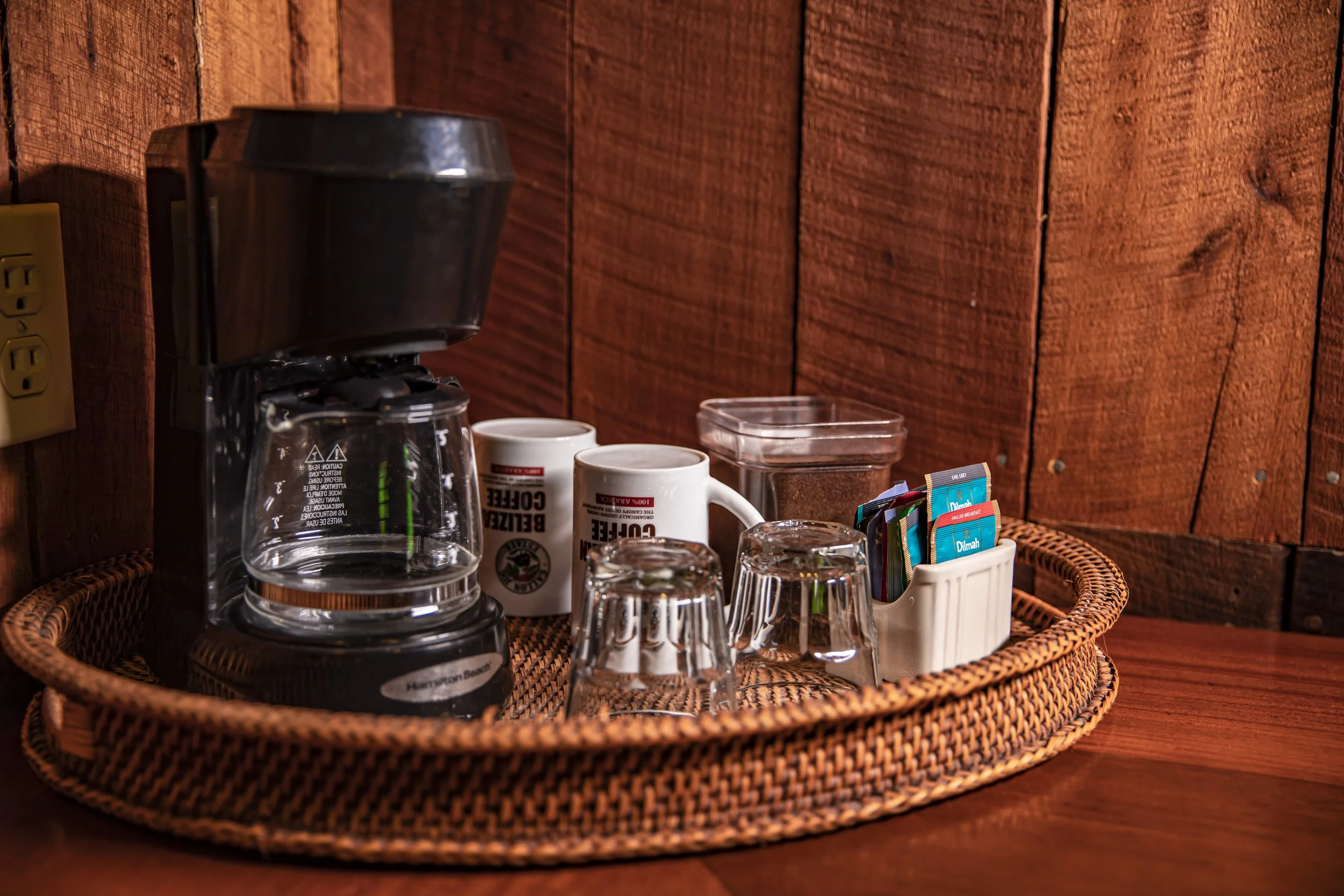 A wicker tray holding a glass coffee carafe, two white mugs, several glasses, and packets of sweeteners on a wooden table against a wooden wall.