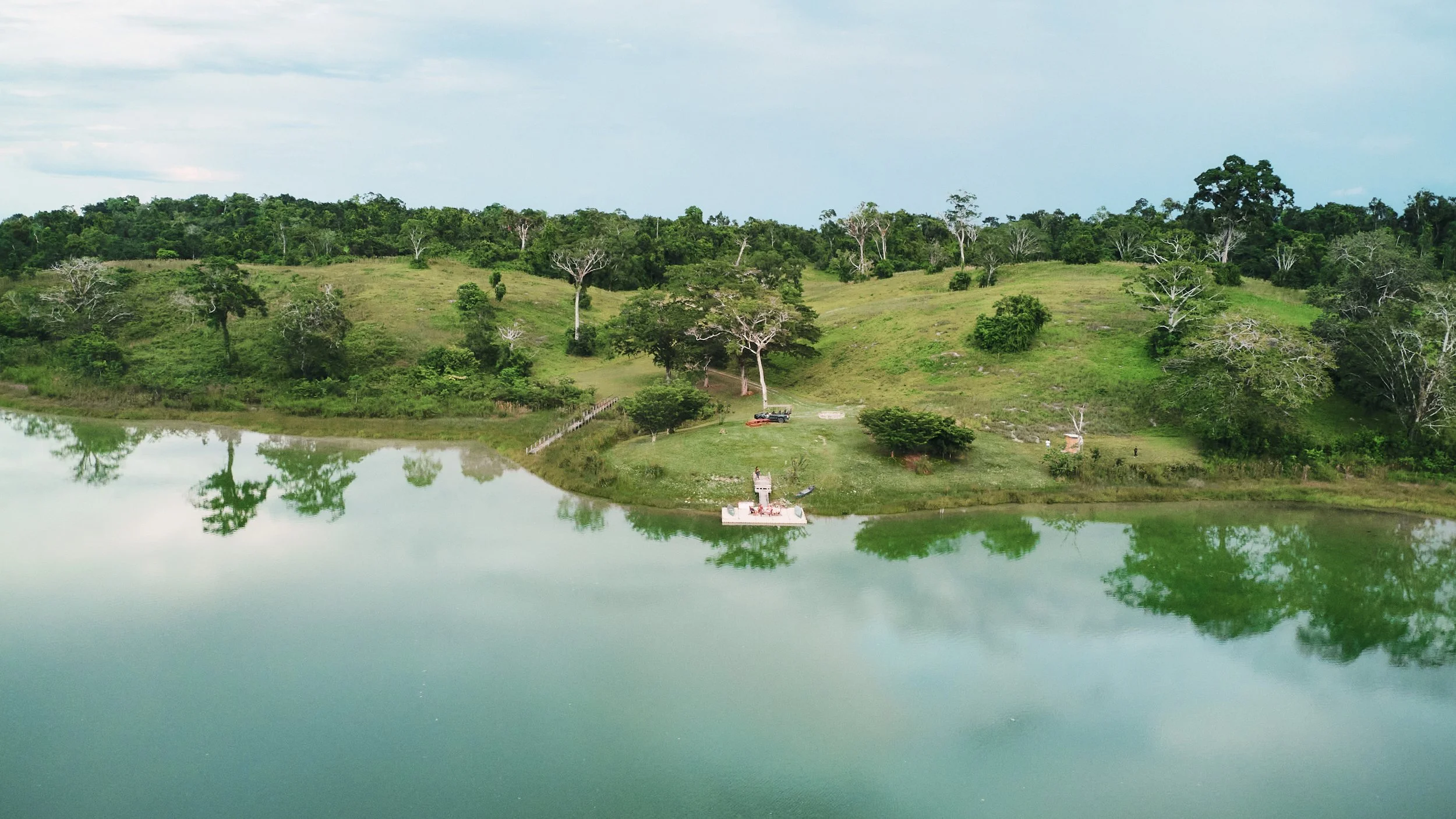 Hillside with scattered trees beside a lagoon, its still water reflecting the trees and cloudy sky
