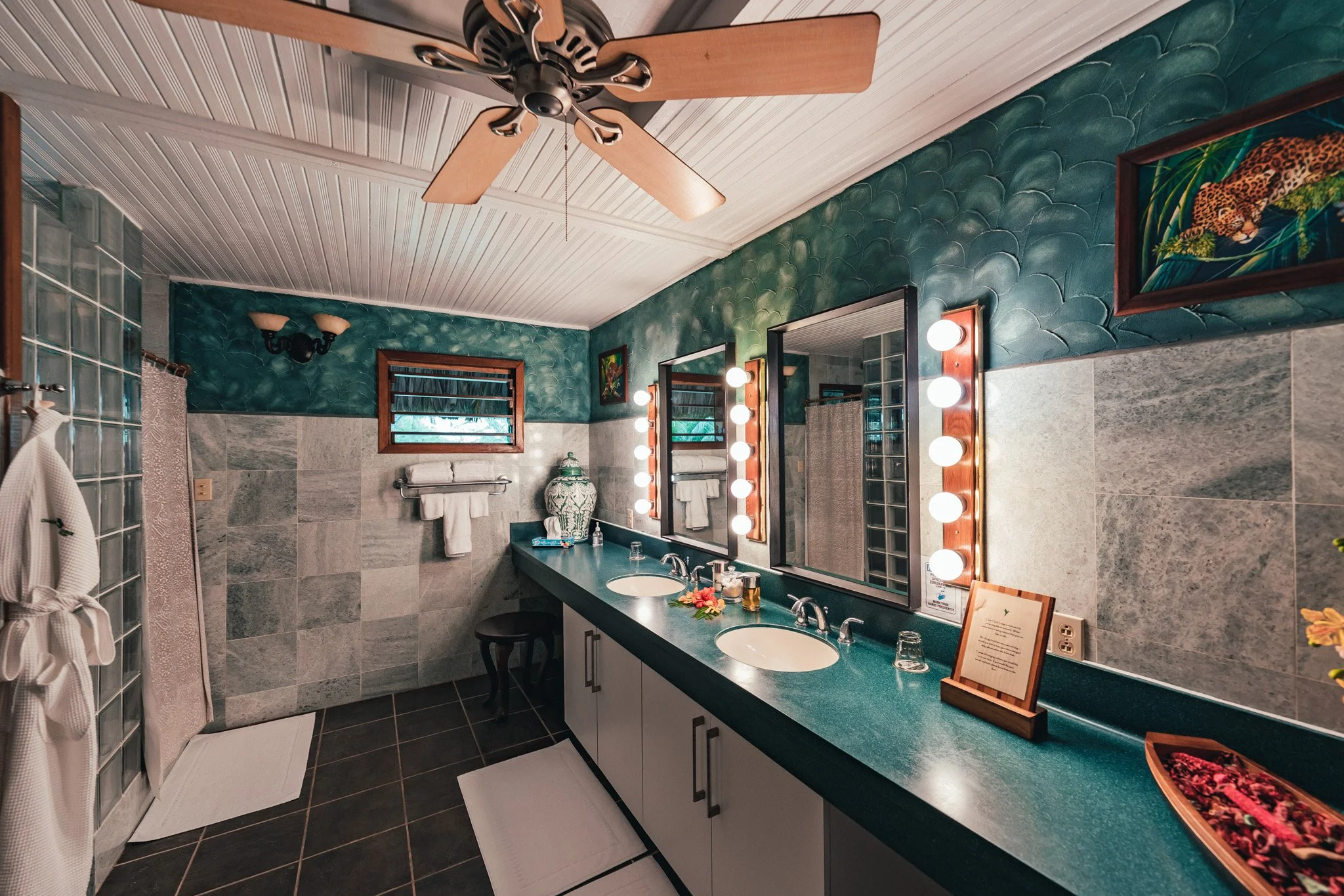 Bathroom with a long green counter, two sinks, a bulb-lit mirror, a teal feature wall and a wood-trimmed window
