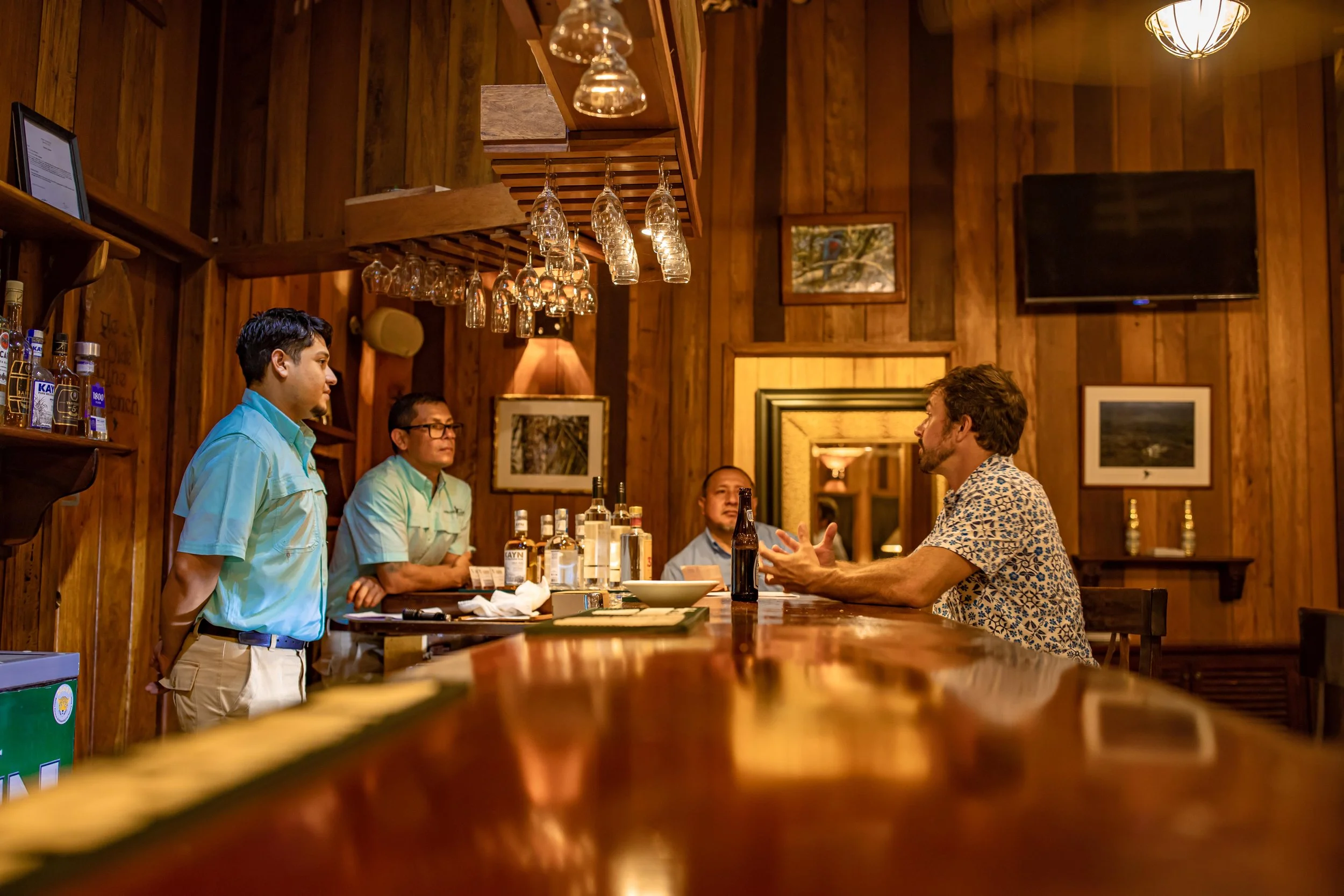 Guests sitting and standing at a wooden bar, talking, in a warmly lit wood-paneled room hung with framed pictures