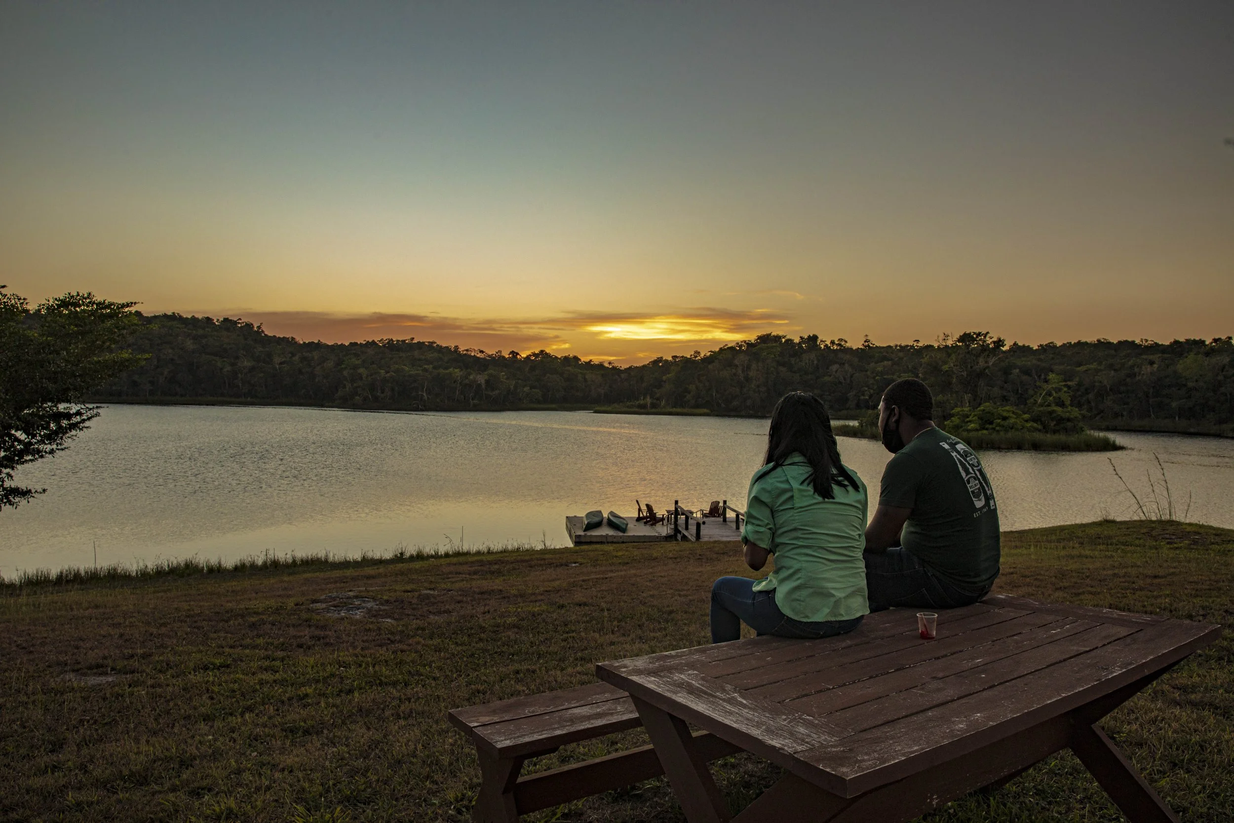 Two people on a bench at the edge of Laguna Verde at sunset, with a small dock and rainforest-covered hills beyond
