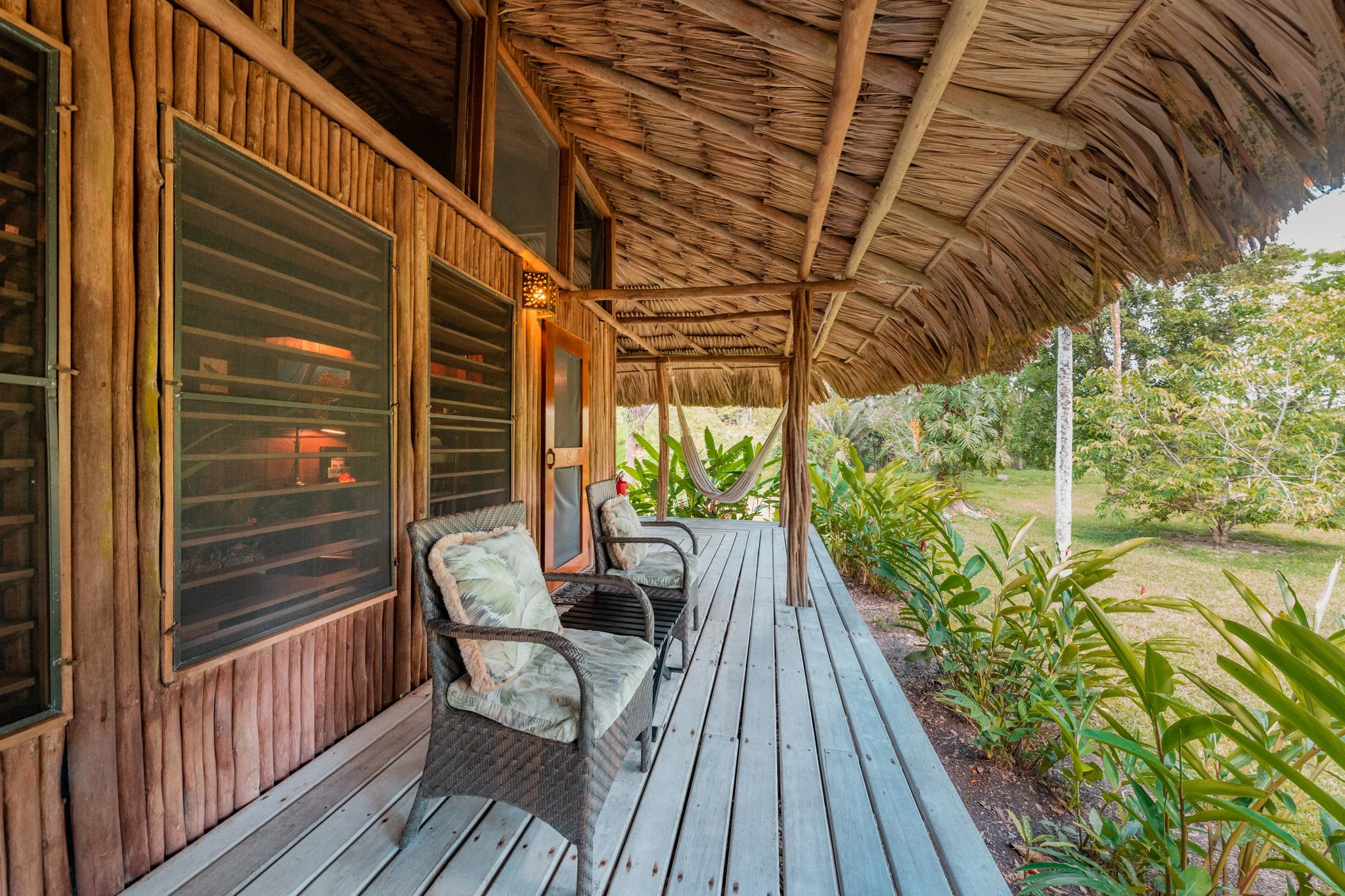 Veranda under a bamboo and thatch roof with three cushioned chairs, facing trees and dense green plants