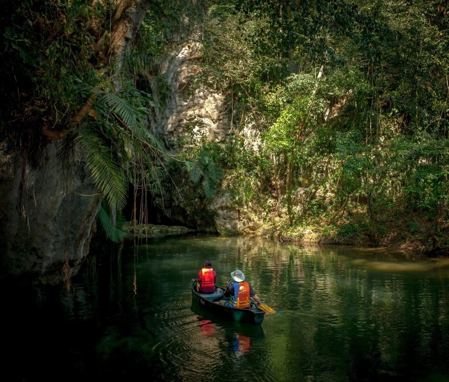 Two people canoeing on a calm river surrounded by dense green jungle, with large rocks and trees overhanging the water.