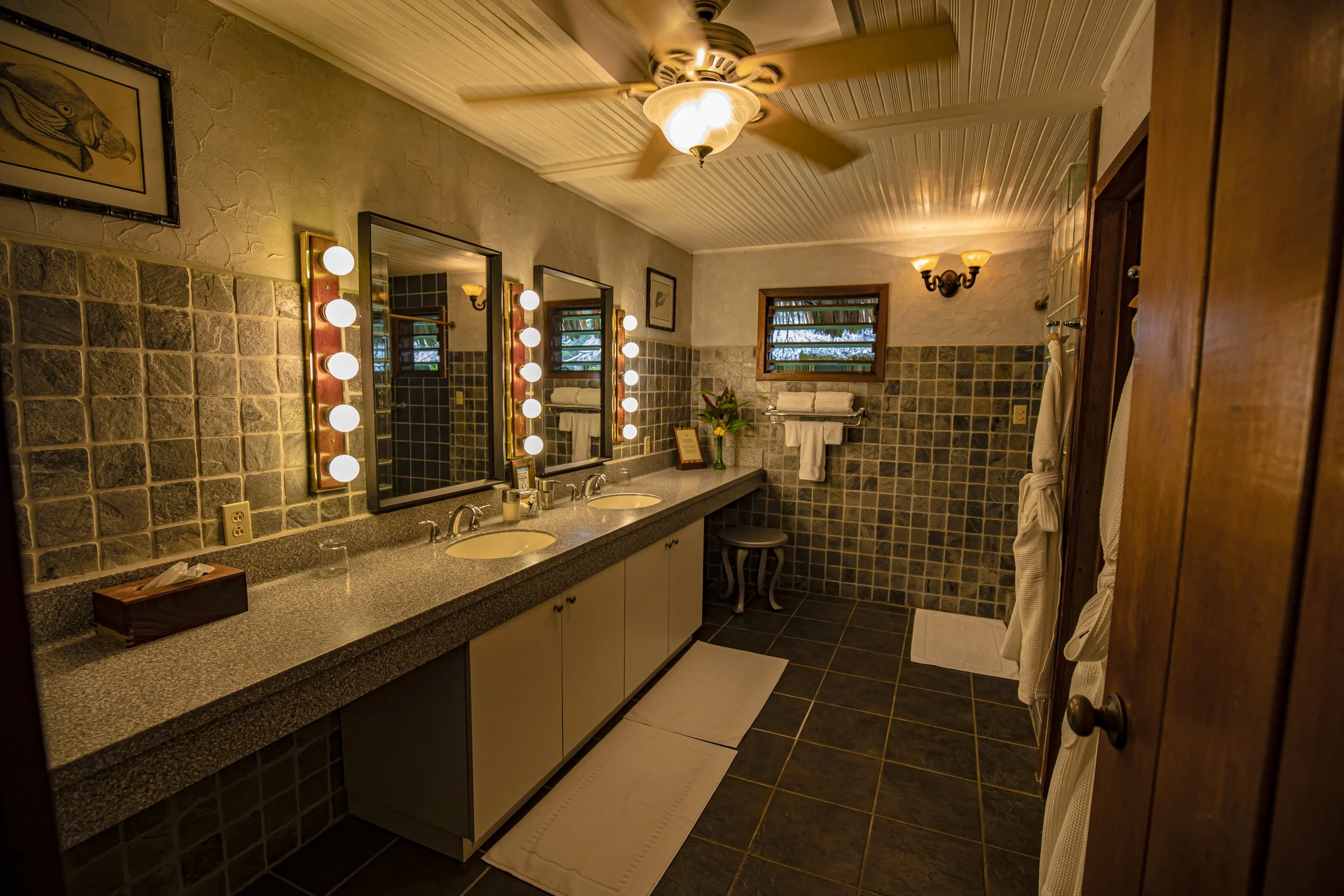 A bathroom with a long countertop featuring two sinks, three mirrors surrounded by light bulbs, a window with blinds, towels hanging on a rack, and a ceiling fan with lights.