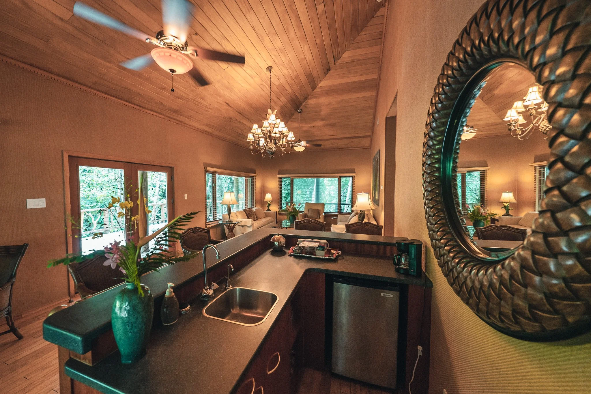 Living room under a high wooden ceiling with a chandelier and ceiling fans, large windows and a counter with a sink