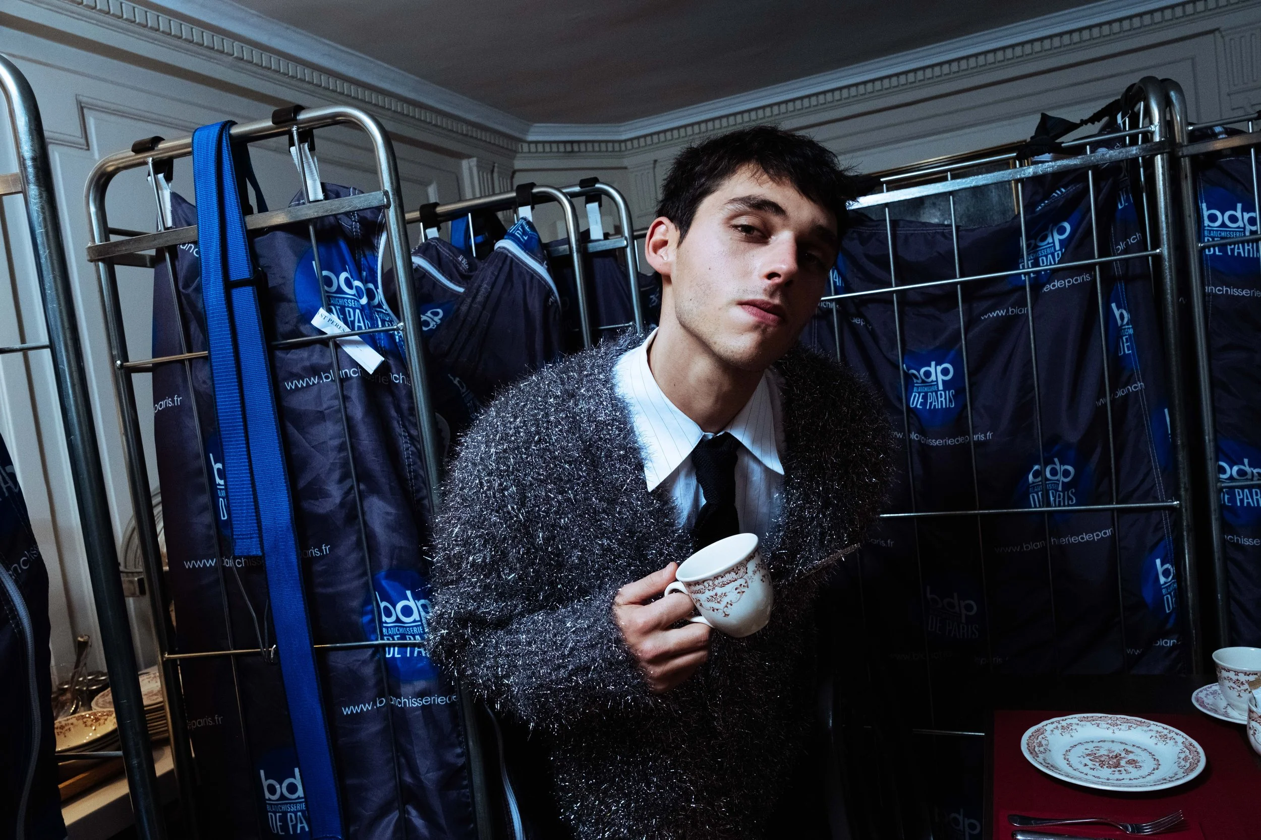 Jeune homme avec des cheveux courts, habillé d'une veste en peluche grise, portant une chemise blanche et une cravate noire, tenant une tasse en porcelaine blanche avec un motif doré, assis devant un présentoir rempli de sacs bleus portant le logo 'bdp' de la blanchisserie de Paris, dans une pièce élégante aux murs ornés de moulures.