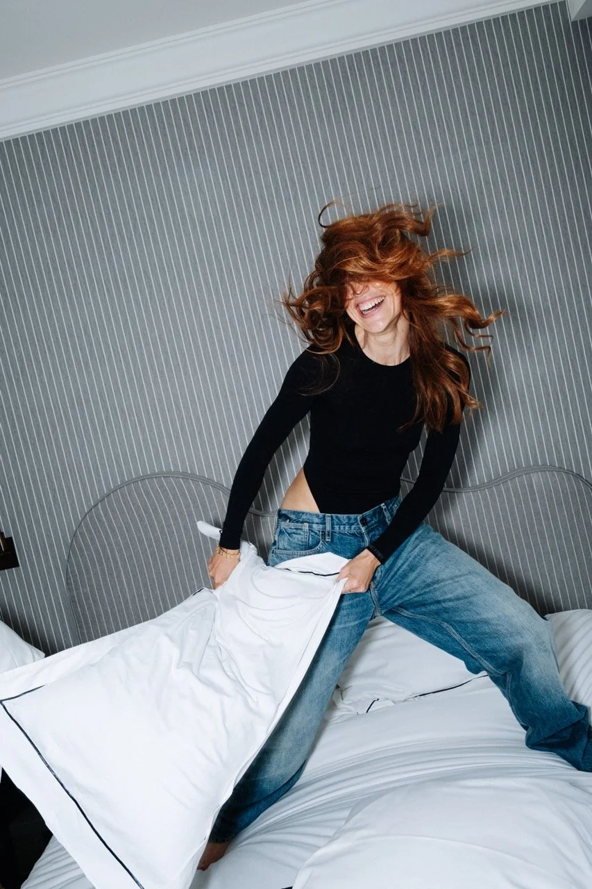 Une femme souriante avec des cheveux bouclés roux, portant un haut noir et un jean, s'amusant en fouettant la couverture d'un lit dans une chambre moderne.
