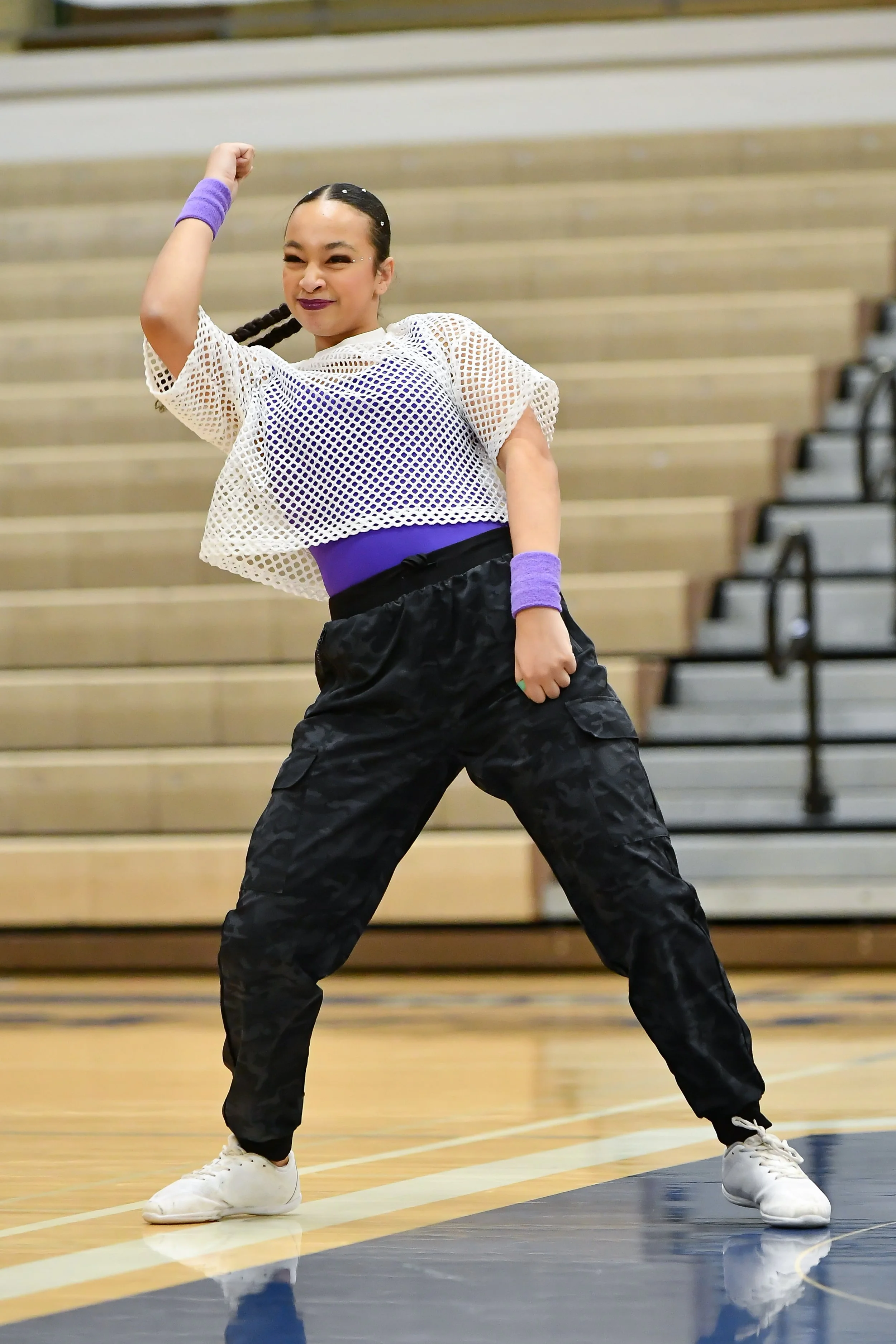 A female dancer performing a dance move in a gymnasium with wooden bleachers in the background. She is wearing a white mesh top, purple sports bra, black pants, and white sneakers, with purple wristbands.